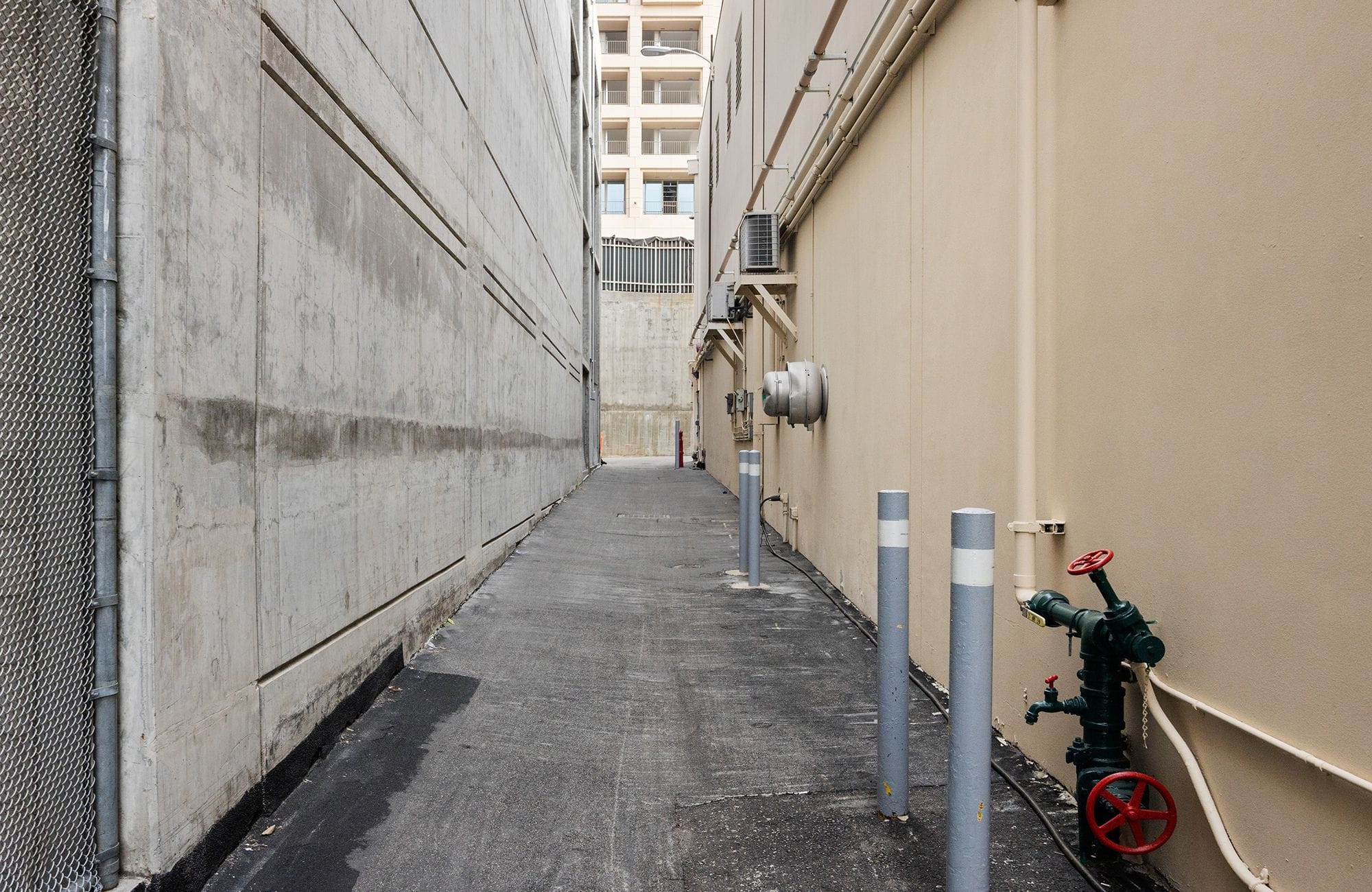 A narrow alley between a concrete wall and a beige wall with a green fire standpipe on the right.