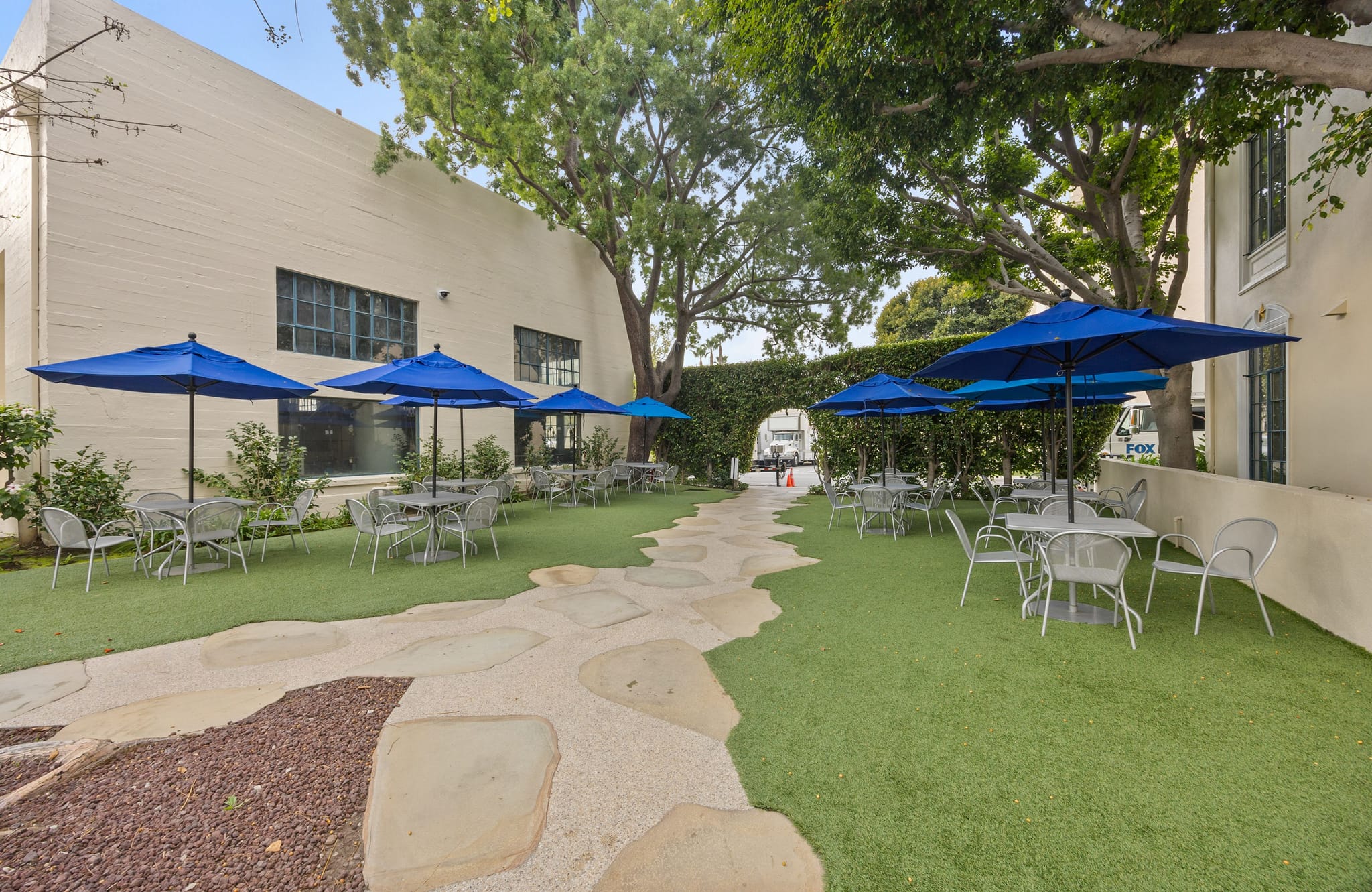 Outdoor patio with green turf, tables, chairs, blue umbrellas, and a winding stone path.