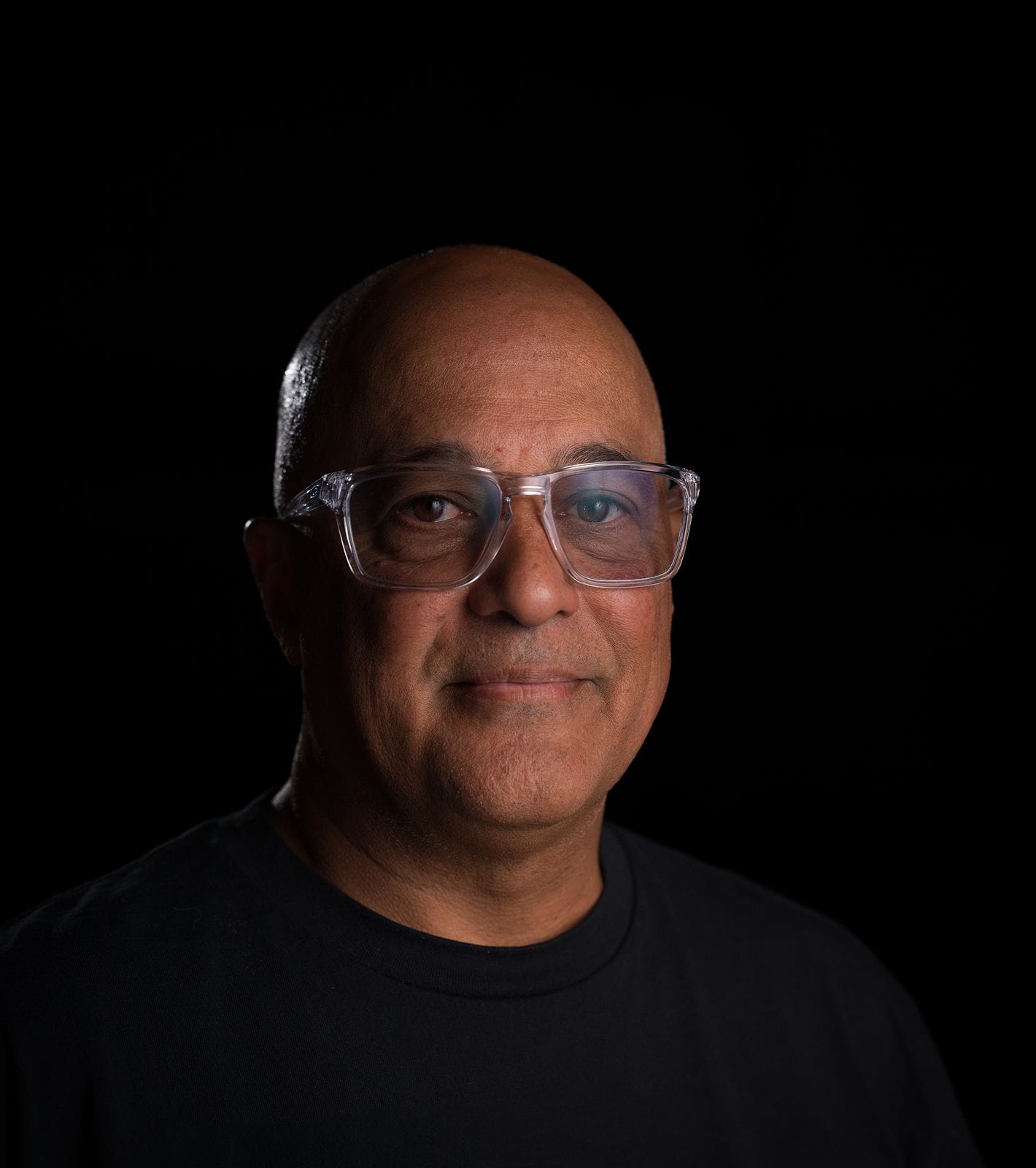 A bald man with clear glasses and a black shirt smiles subtly against a black background.
