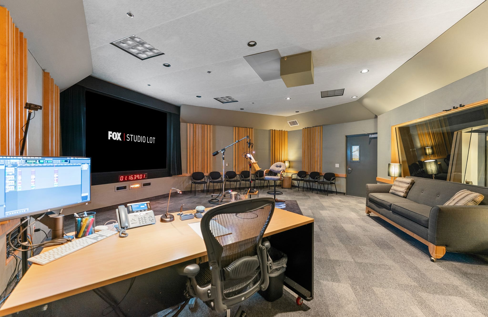 A large sound studio with a control desk, a screen displaying "FOX | STUDIO LOT", microphones, and a gray sofa.