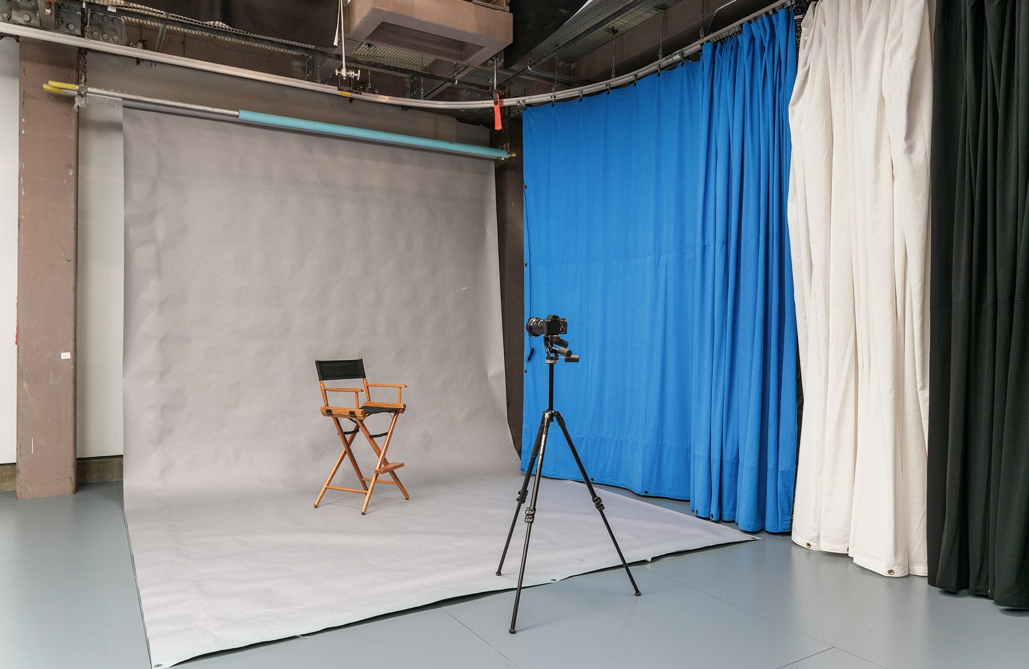 A photography studio with a gray seamless backdrop, a director's chair, a camera on a tripod, and blue, white, and black curtains.