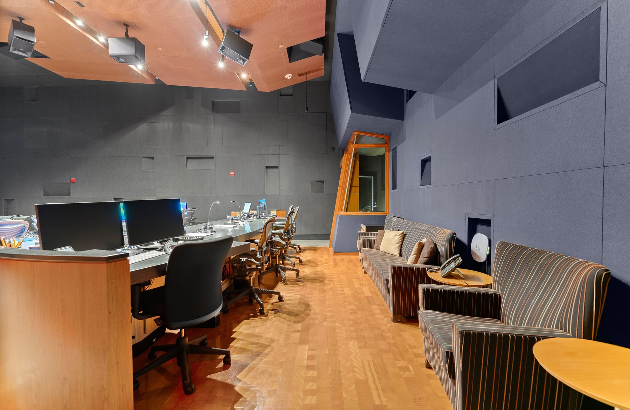 Modern studio interior with a control desk and monitors on the left, and a lounge area with striped sofas on the right.