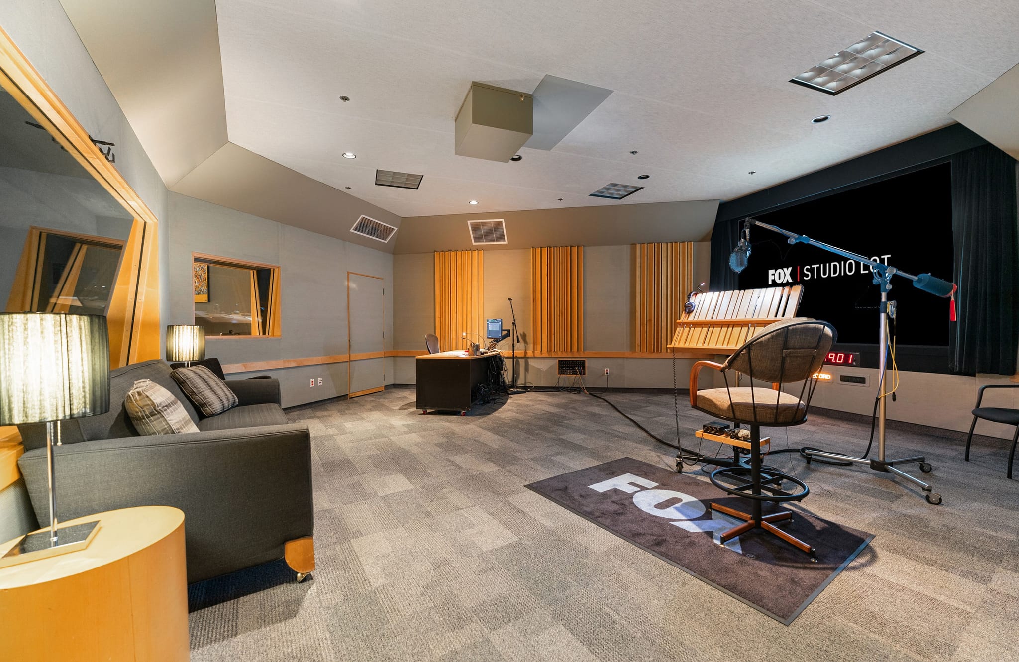 A professional sound studio with a large screen displaying "FOX STUDIO LOT", a microphone, acoustic panels, and a sofa.
