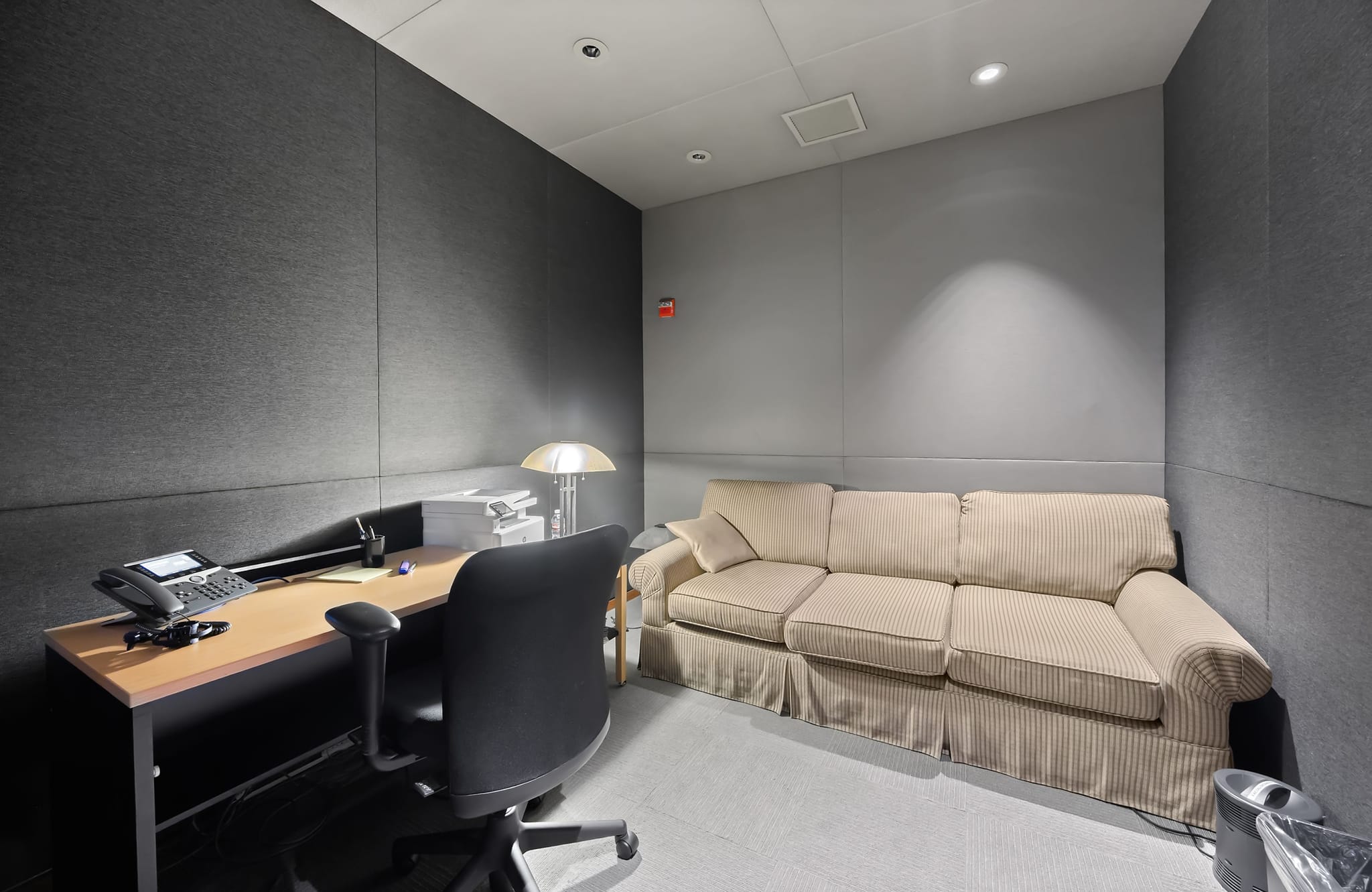A small room with a desk, chair, and phone on the left, and a beige three-seater sofa on the right, all surrounded by gray acoustic wall panels.