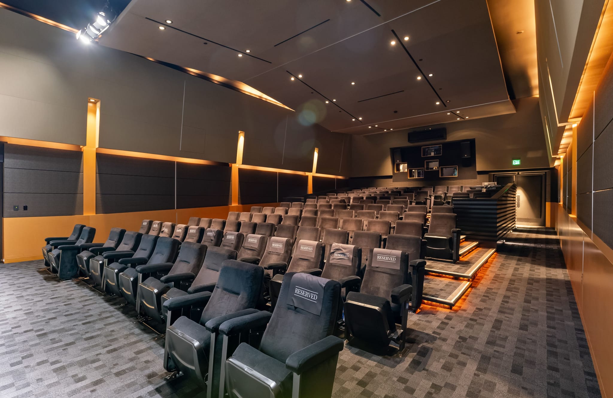 Empty modern screening room with rows of dark recliners, warm accent lighting, and orange wall panels.