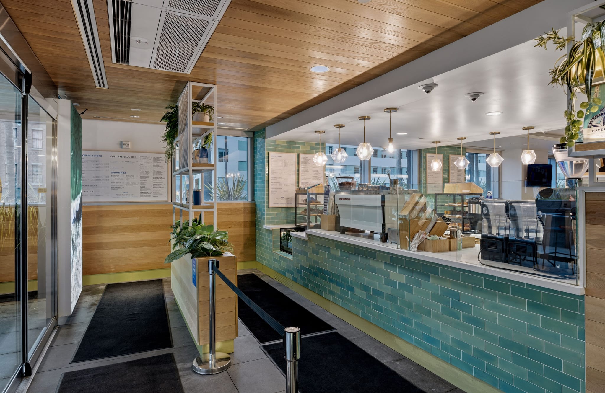 A bright, modern cafe interior with a teal-tiled counter, coffee machines, light wood paneling, and plants.