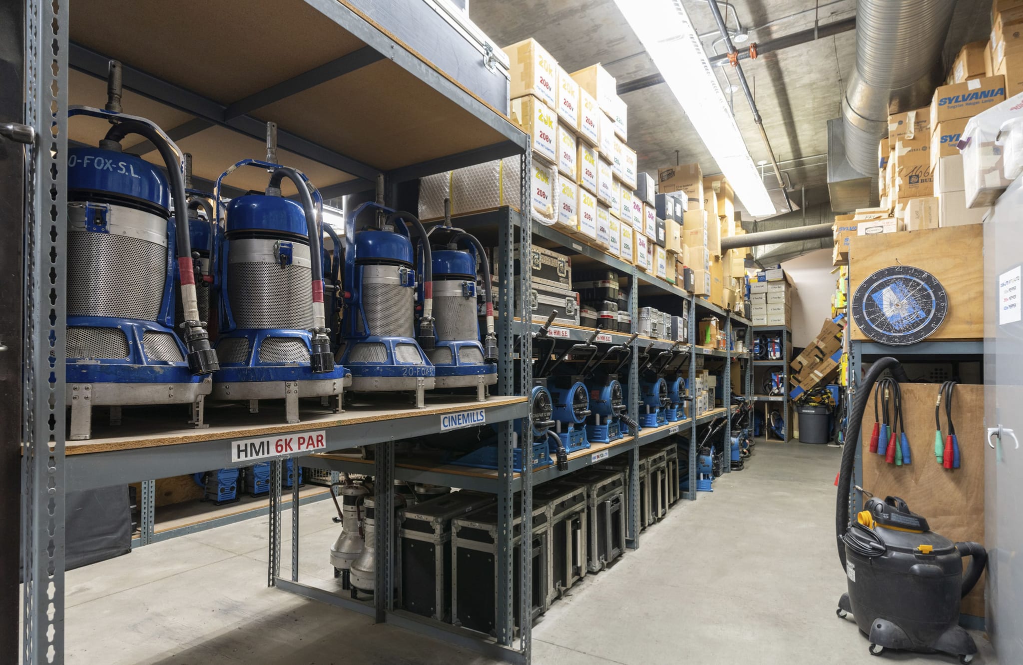 Storage room with metal shelves filled with HMI lights, other equipment, and cardboard boxes.