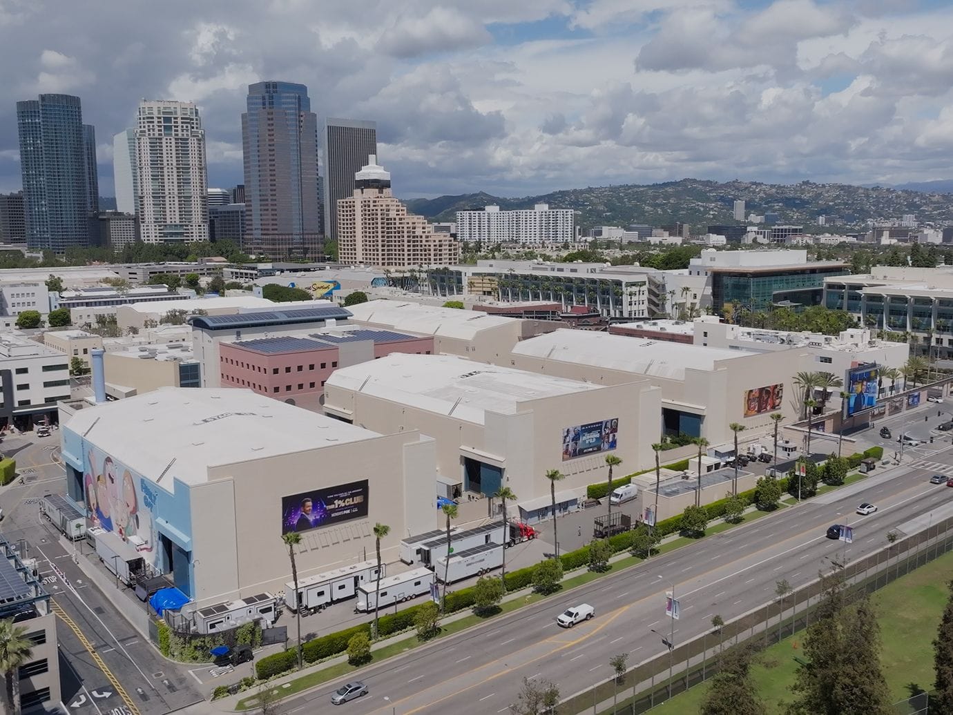 Aerial view of a large studio complex alongside a busy road, with a city skyline and hills in the background.