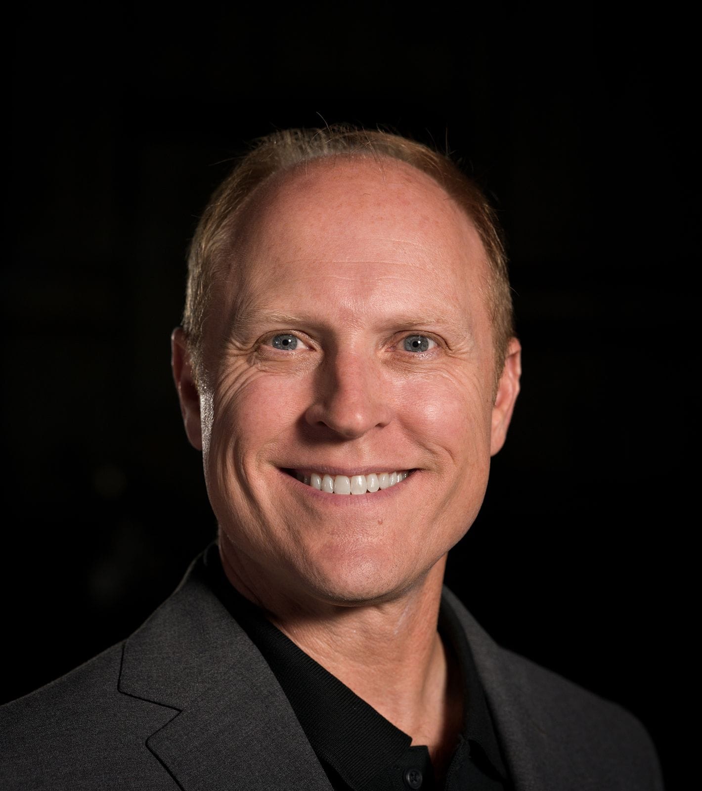 Smiling man with light brown hair, blue eyes, and a dark collared shirt.