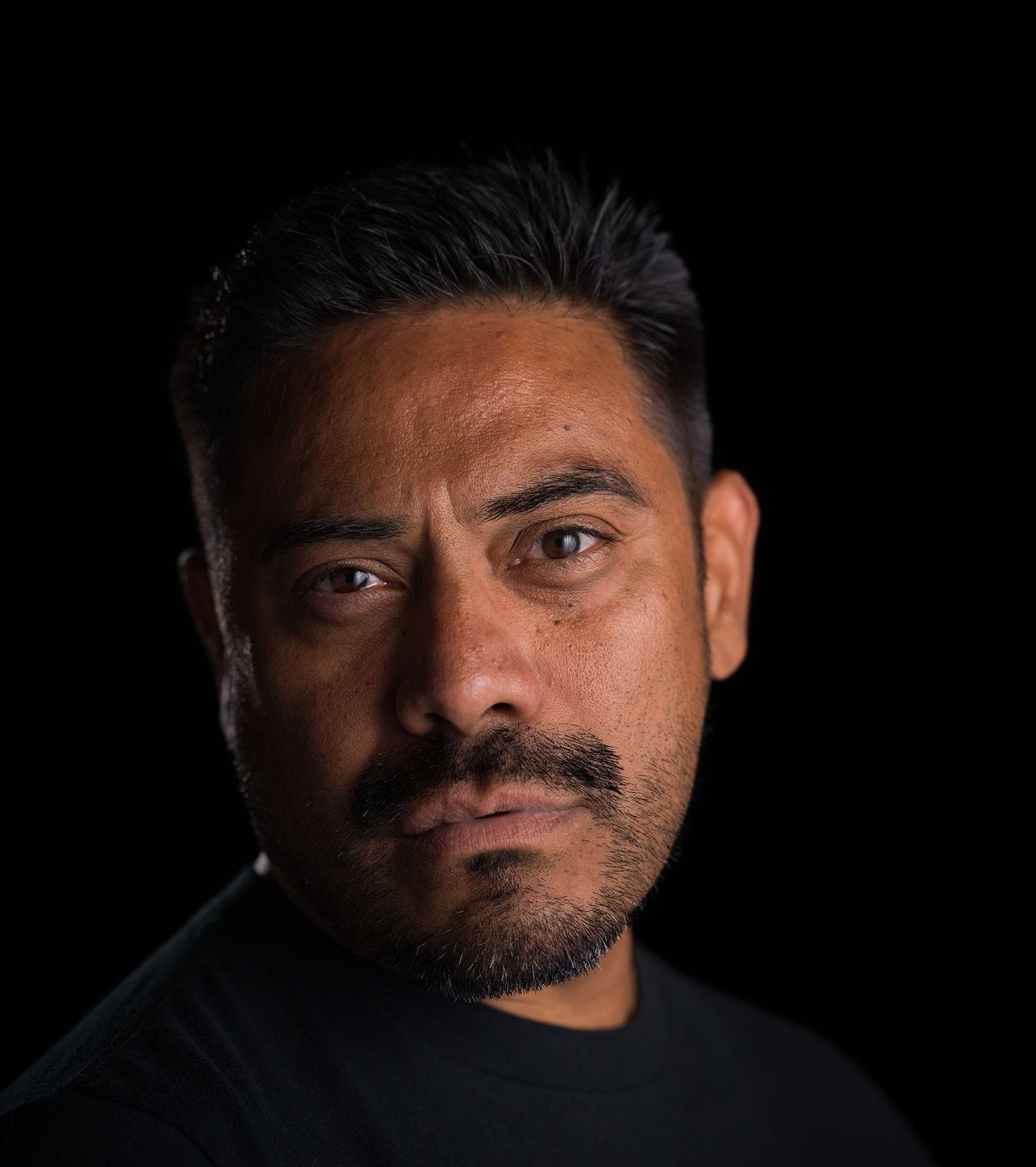 Portrait of a man with dark hair and a beard, looking directly forward against a dark background.