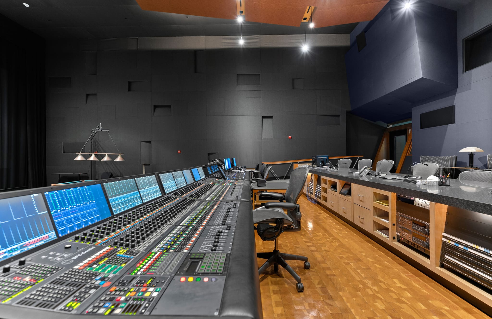 A modern audio control room featuring a large mixing console with multiple screens, dark acoustically treated walls, and a wooden floor.