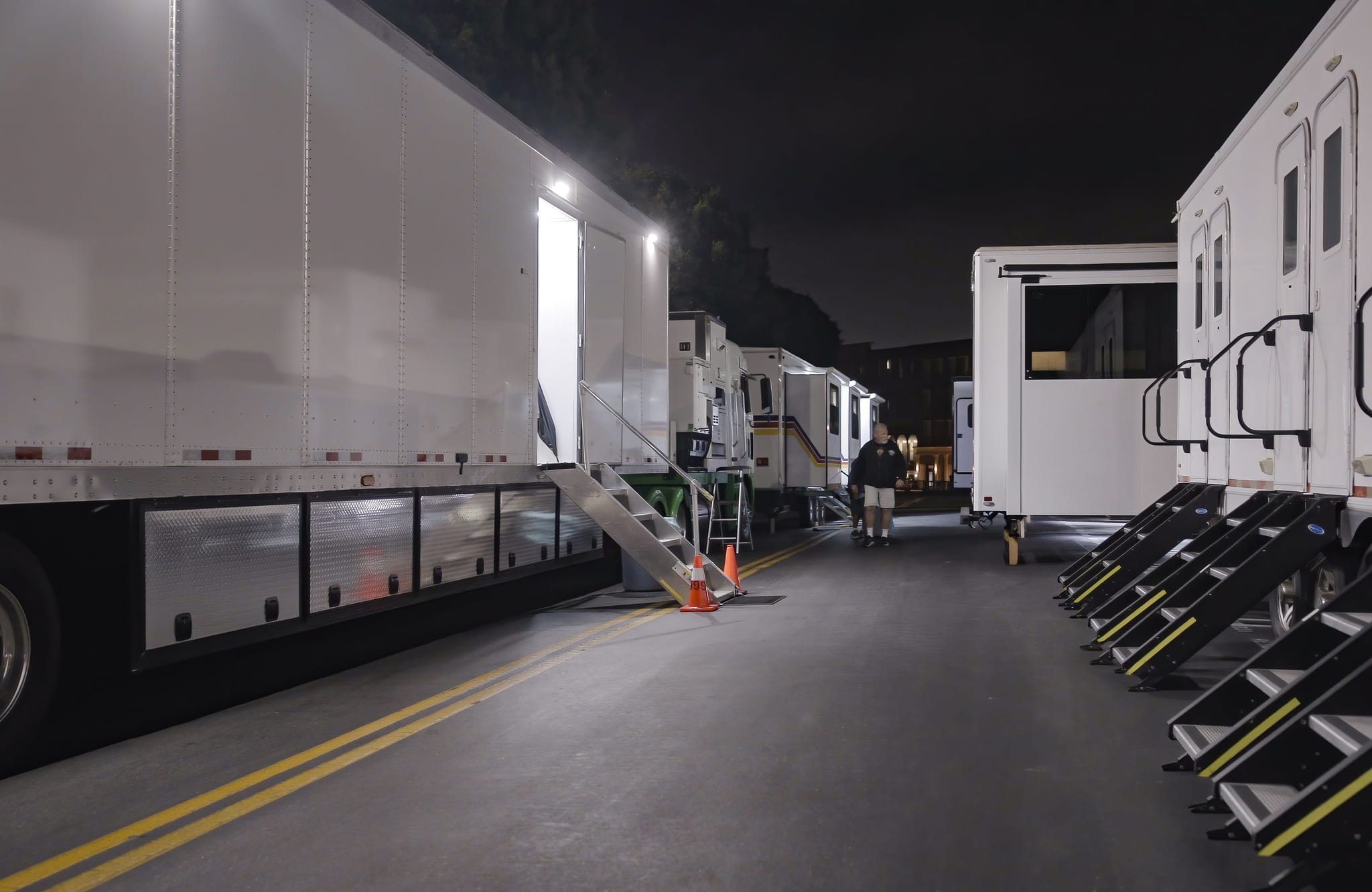 Film set trailers parked along a street at night, with a person walking in the distance.
