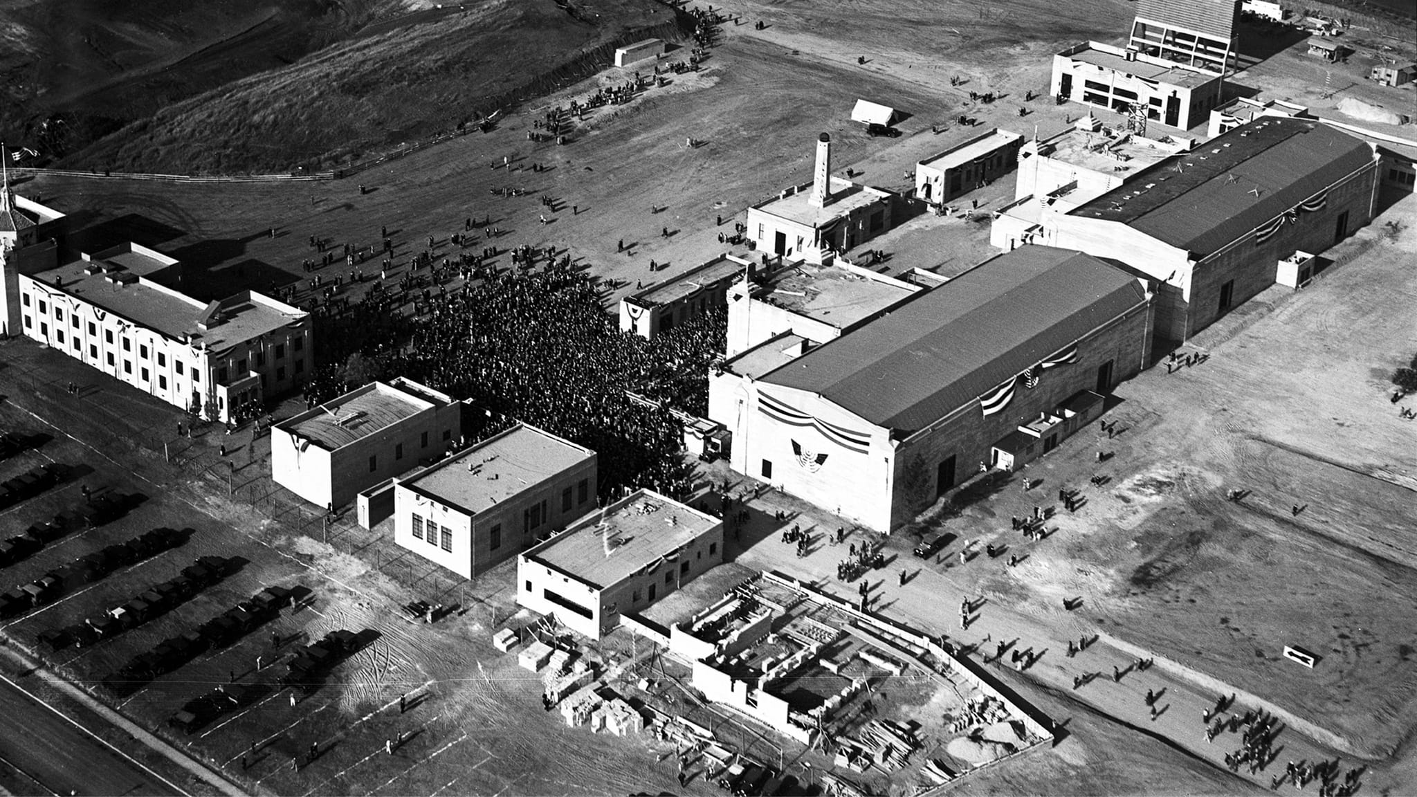 Aerial black and white photo of a large building complex adorned with banners, surrounded by many people.