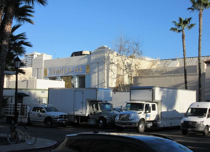 White trucks, some with FOX logos, parked on a street in front of a building labeled "STAGES 1 & 2" with palm trees and a clear blue sky.