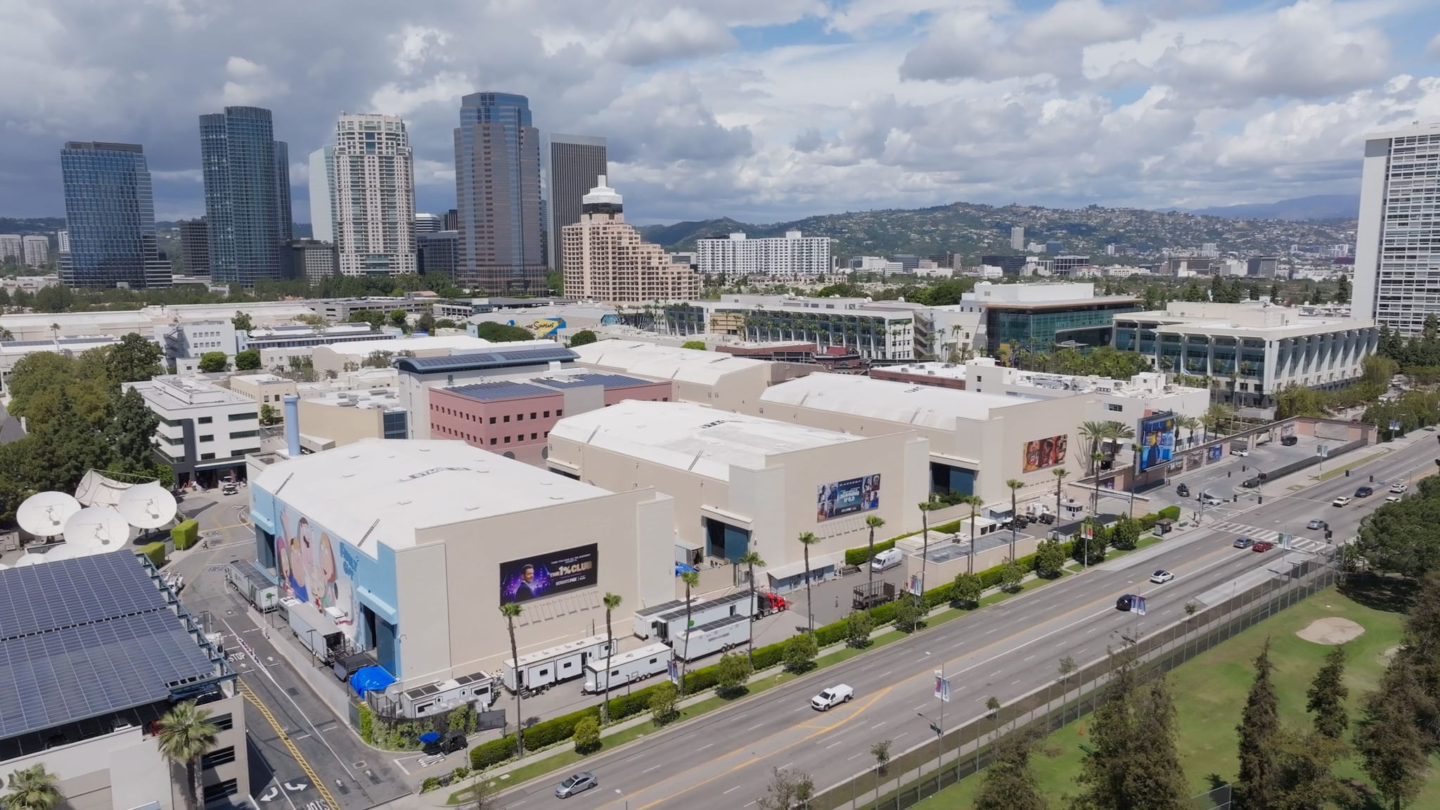 Aerial view of a large film studio complex with promotional banners, a multi-lane road, and a distant city skyline with hills.