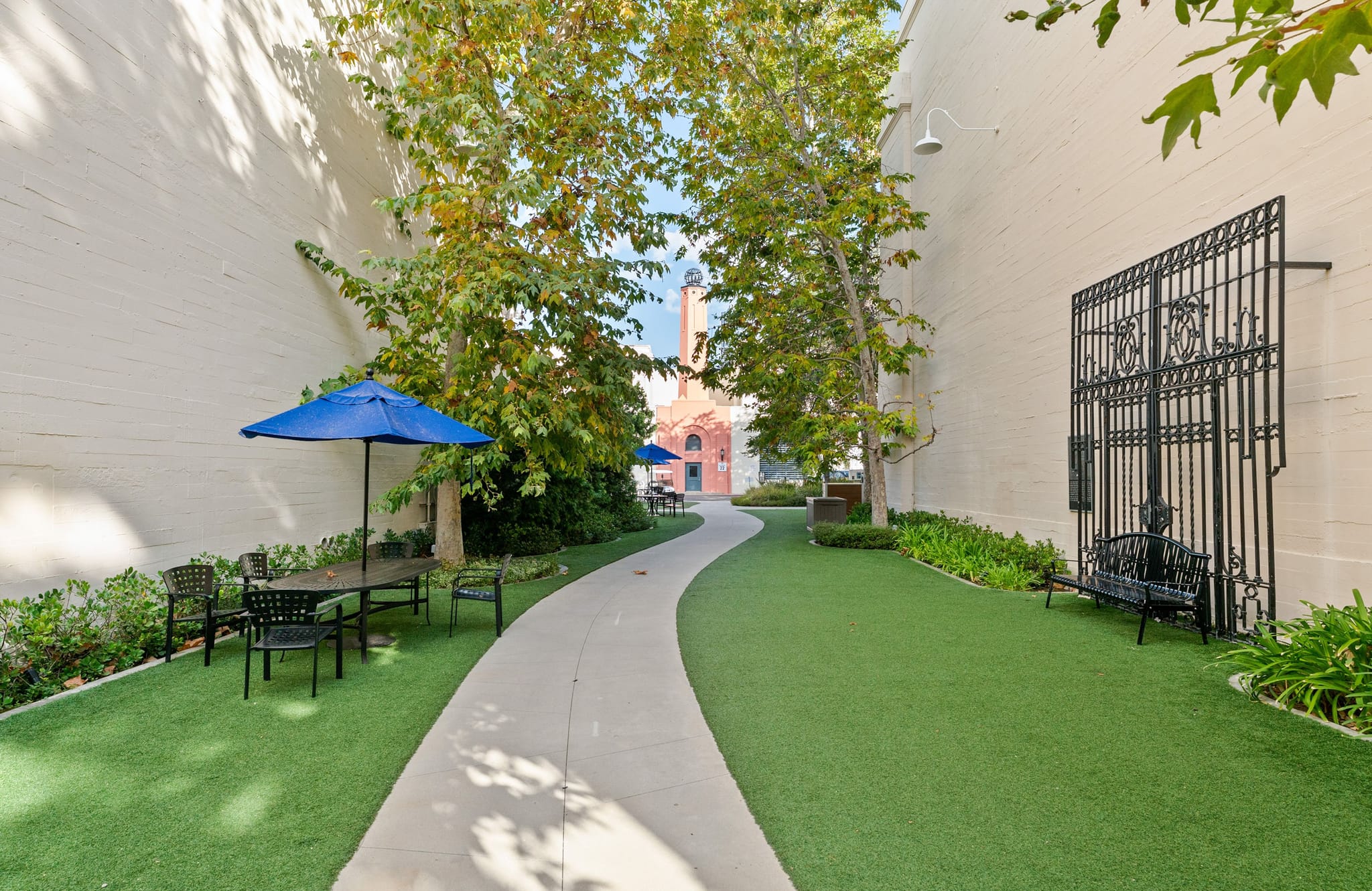 A long, narrow courtyard with a winding path on artificial turf, a blue umbrella over a dining set, a decorative black gate, and a brick building in the background.