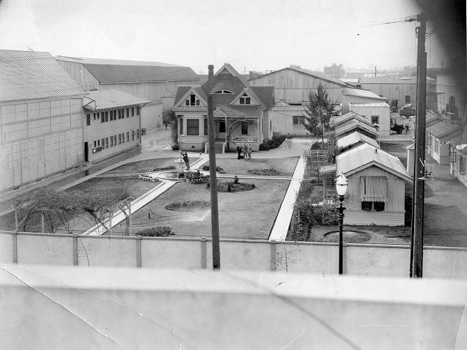 Elevated black and white view of a house with a garden, surrounded by smaller bungalows and large warehouse-like buildings.