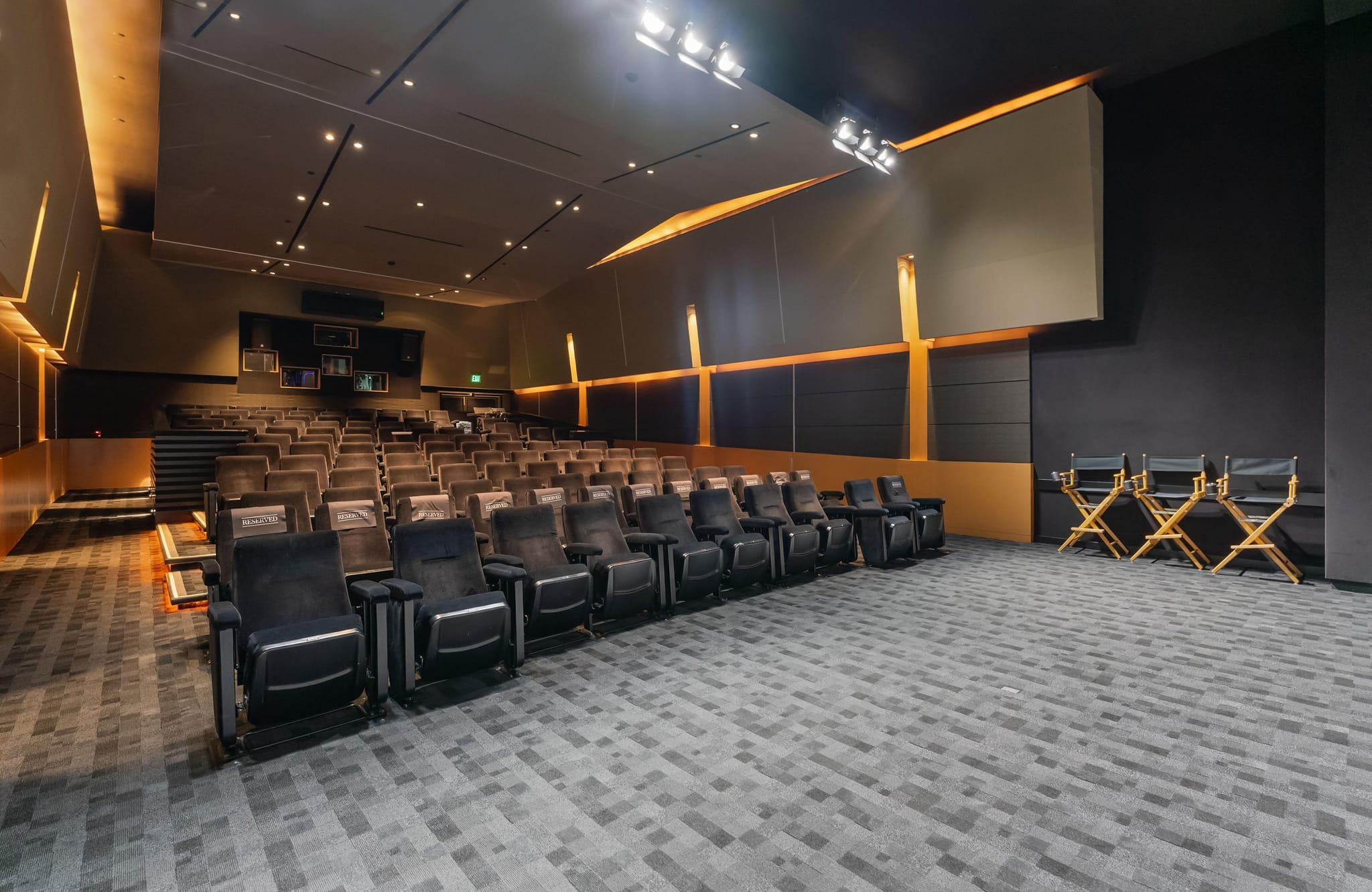 A modern screening room with rows of dark theater seats, a large screen, accent lighting, and three director's chairs.