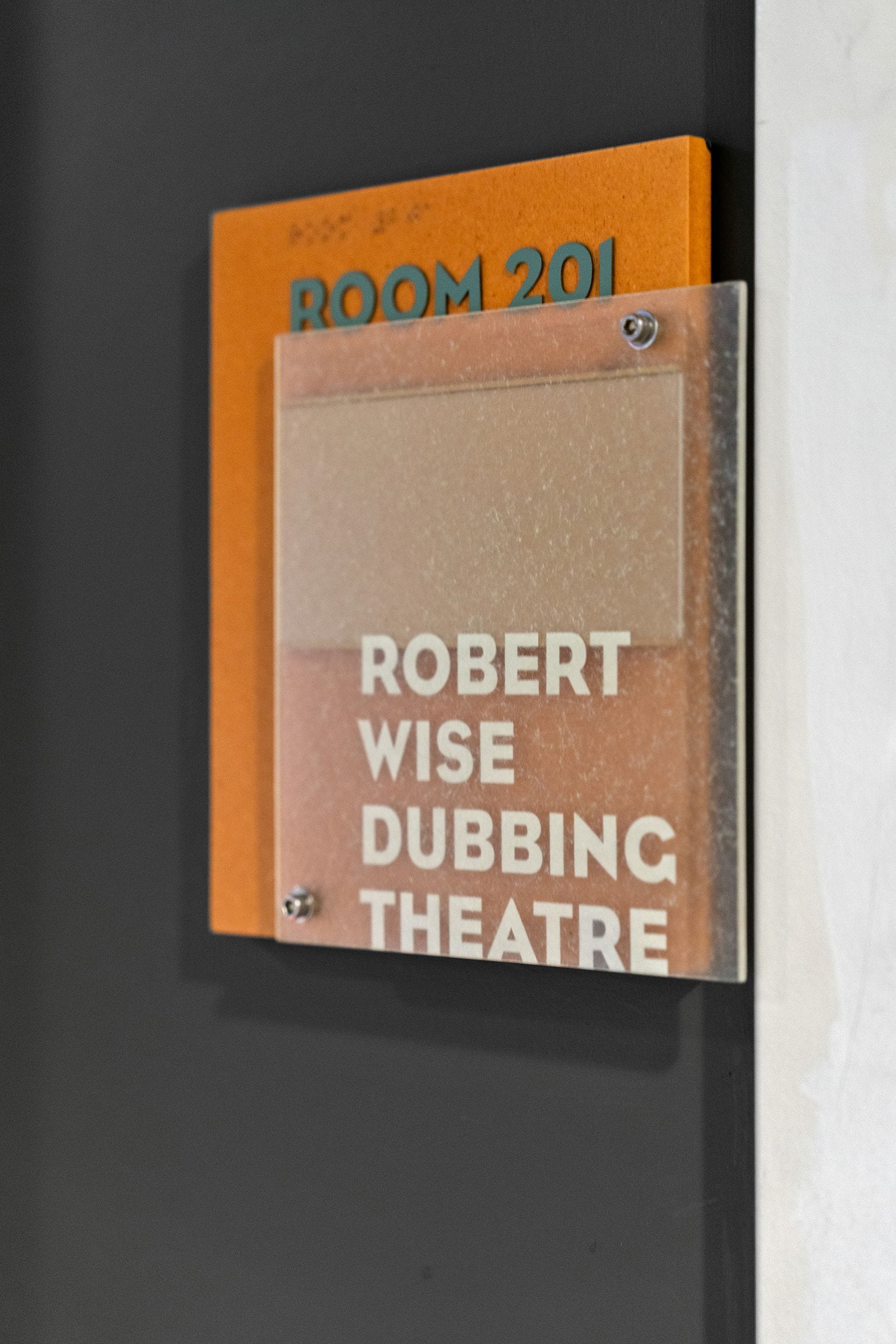 An orange sign for "ROOM 201" with braille, layered with a frosted acrylic panel displaying "ROBERT WISE DUBBING THEATRE", on a dark wall.