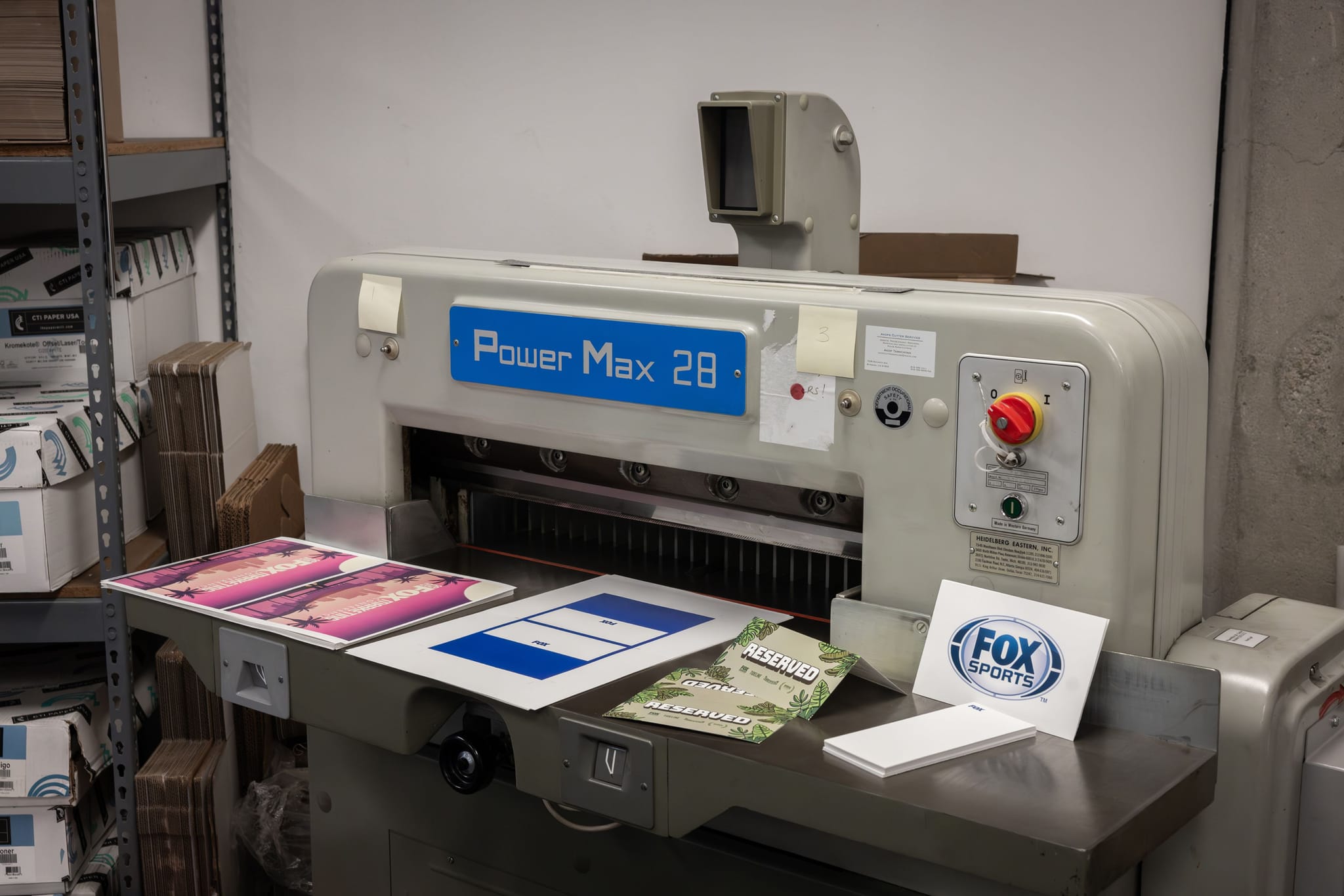 Power Max 28 industrial paper cutter with printed materials like Fox Sports cards and reserved signs on its surface.