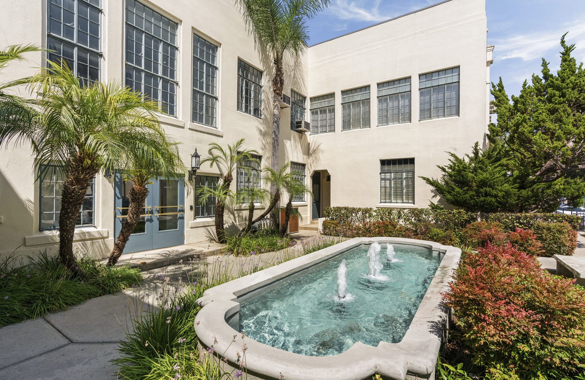 A courtyard with a rectangular fountain, palm trees, lush plants, and a light-colored building.
