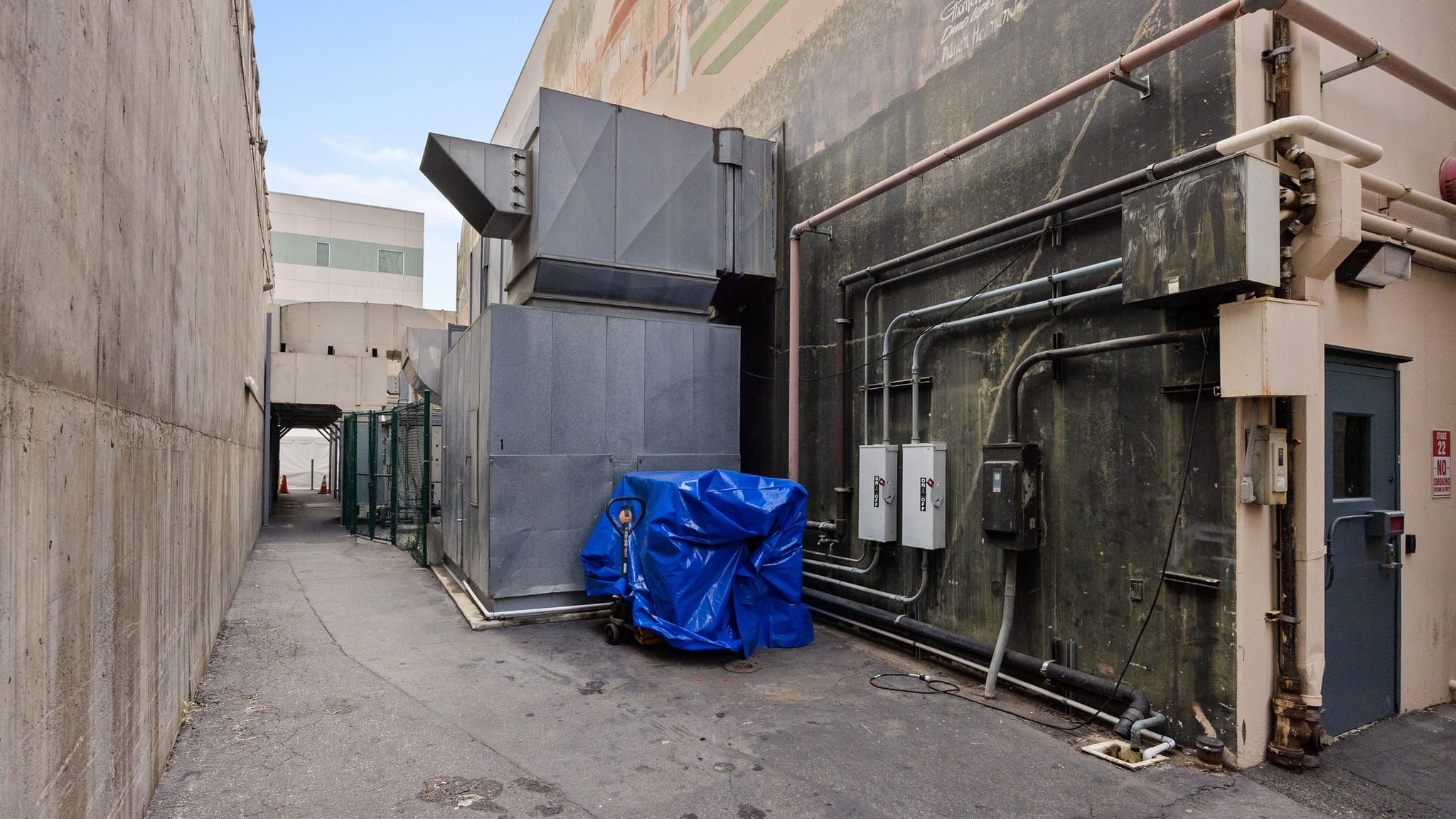 Narrow industrial alley between a concrete wall and a building with HVAC units and utility pipes.