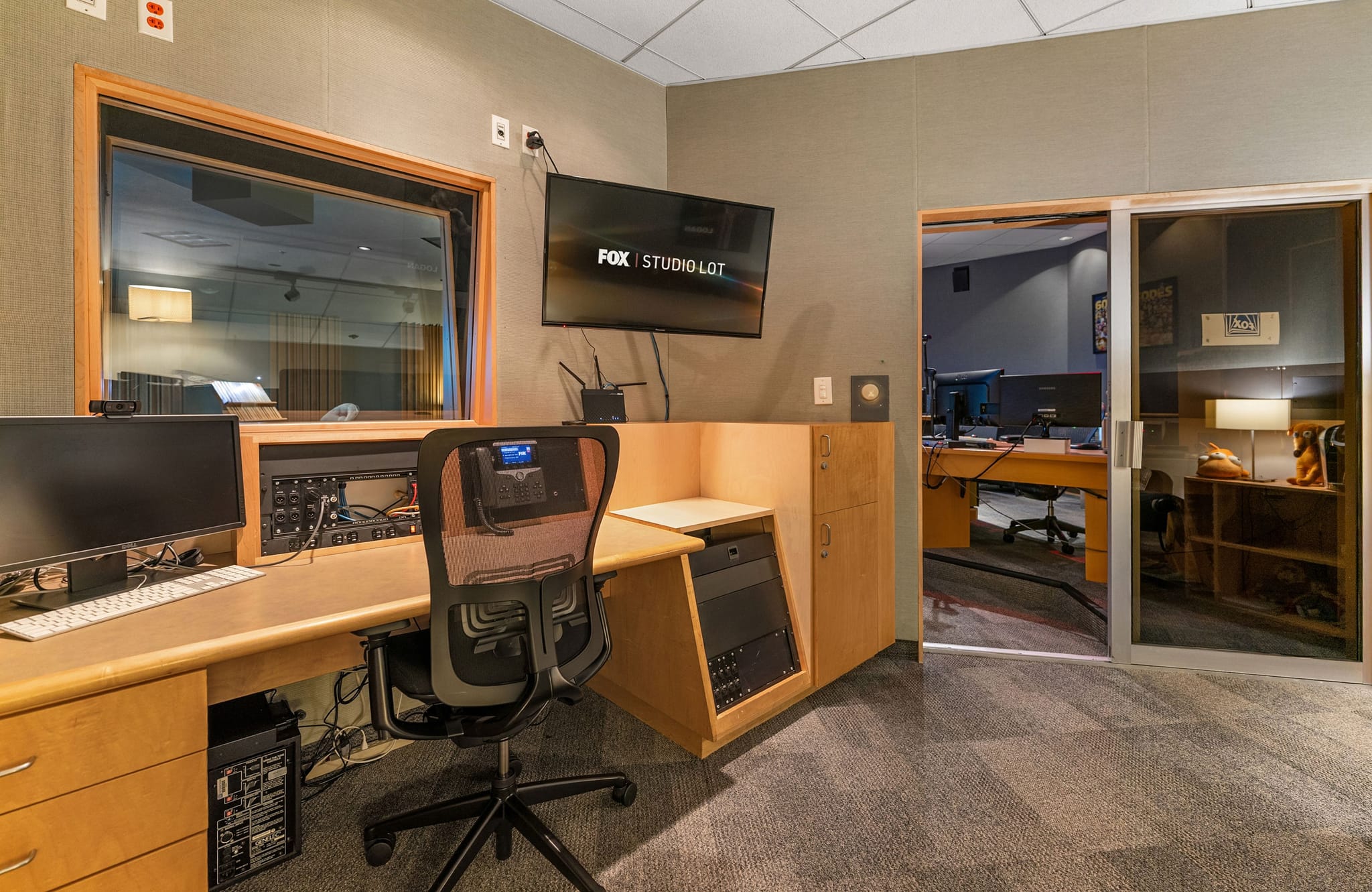 A recording studio control room with a desk, monitors, soundboard, a window into a sound booth, and a TV displaying "FOX | STUDIO LOT".