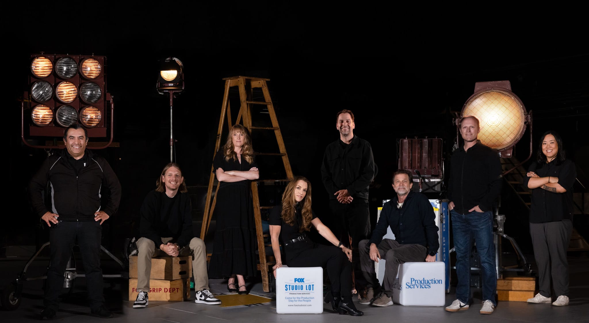 Eight production team members, four men and four women, pose in a studio with large lights and a ladder against a dark background. Two are seated on boxes labeled "FOX STUDIO LOT" and "Production Services."