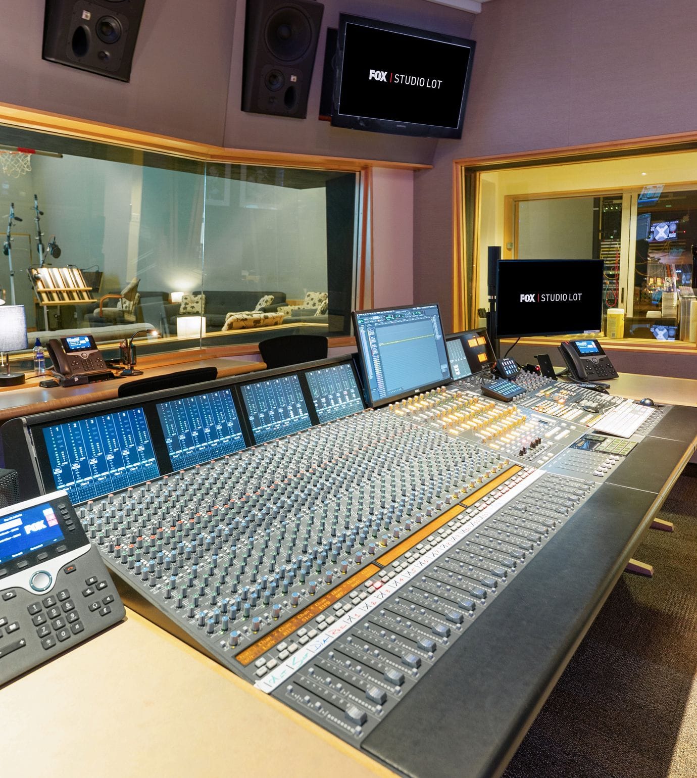 Professional audio mixing console in a studio control room, with a recording booth through glass and a monitor showing 'FOX | STUDIO LOT'.