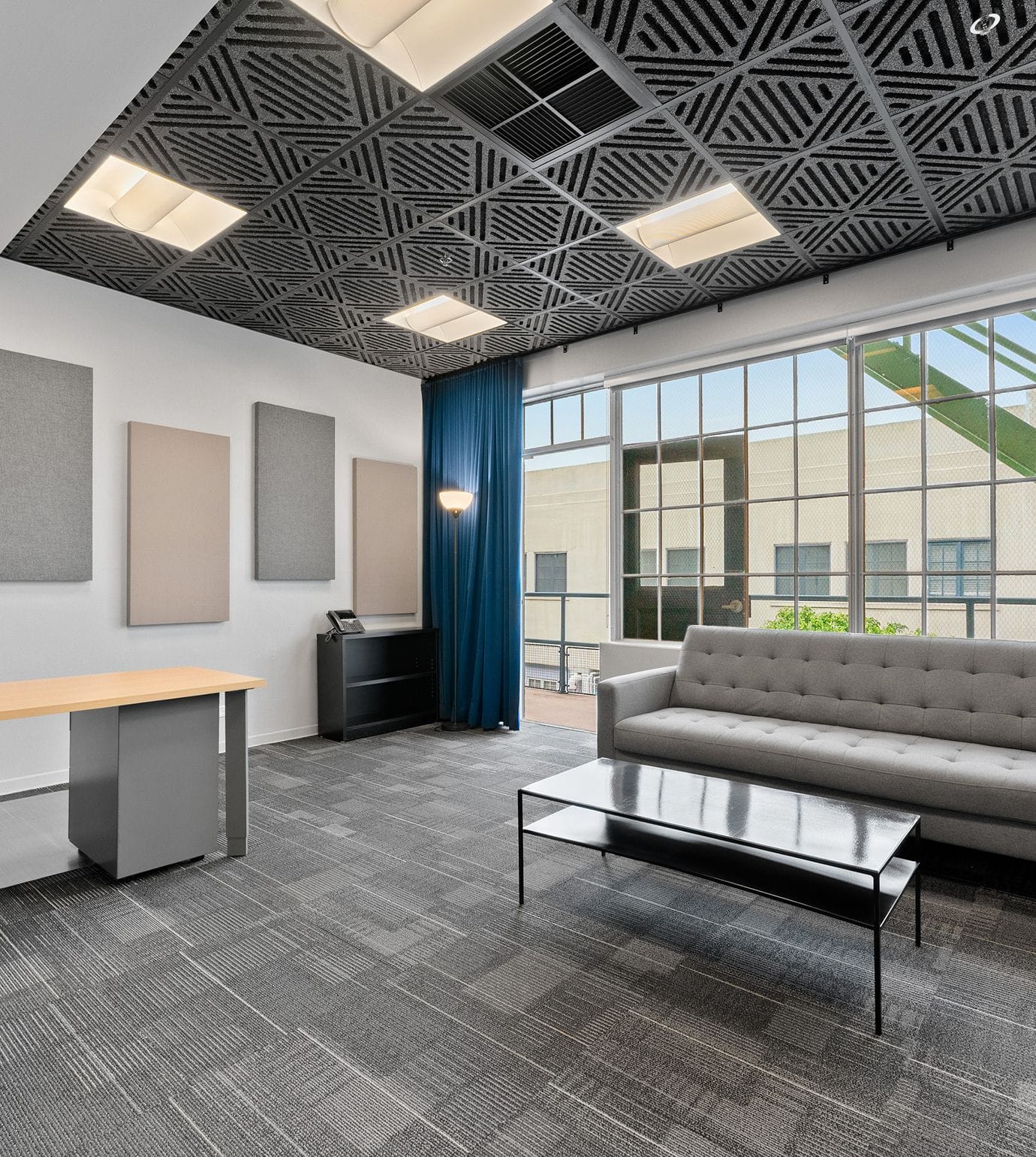 A modern office lounge with a dark patterned ceiling, large windows, grey sofa, black coffee table, light wood desk, and acoustic wall panels.