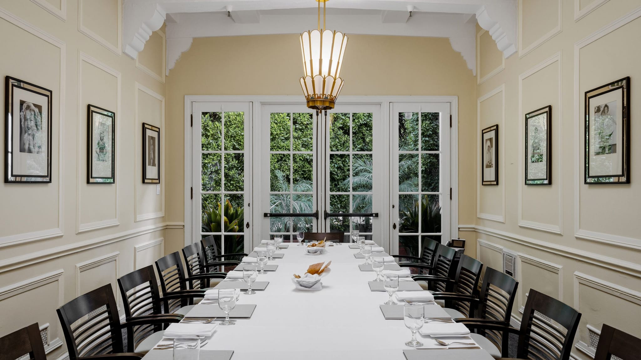 A formal dining or meeting room with a long table set for many guests, French doors overlooking greenery, and a decorative chandelier.