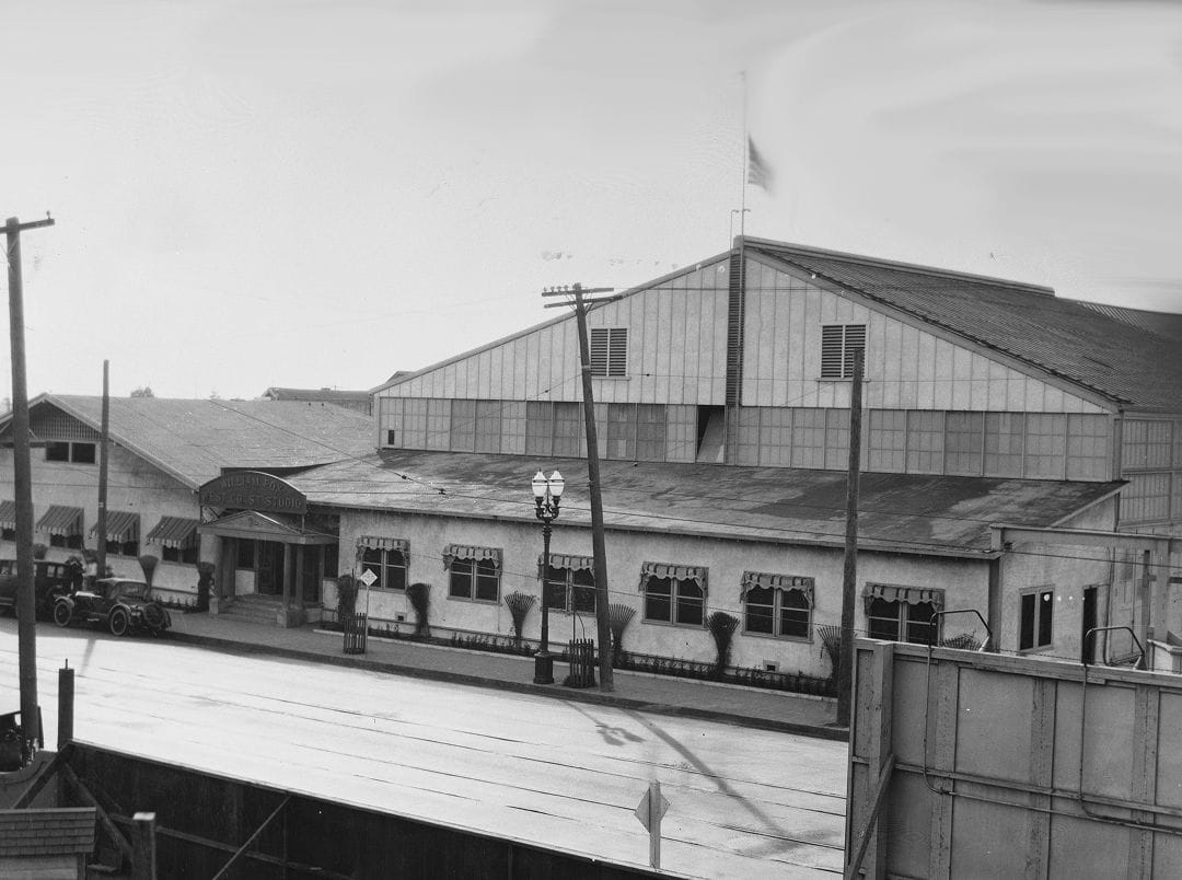 Vintage black and white photo of an industrial street scene, featuring a large hangar, smaller buildings, and vintage cars.