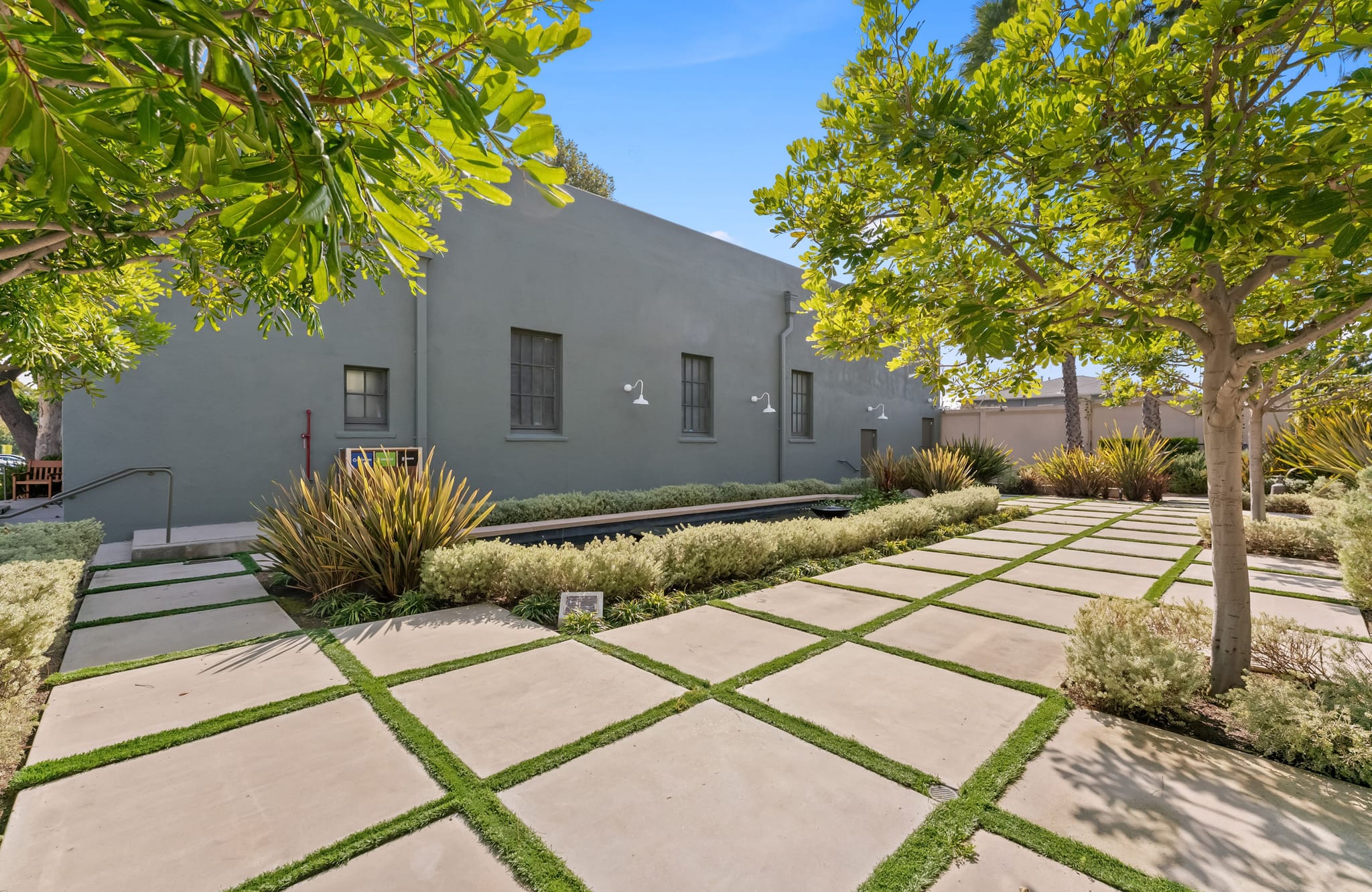 Modern outdoor courtyard with concrete pavers and grass strips, leading to a grey building, surrounded by trees and shrubs under a blue sky.