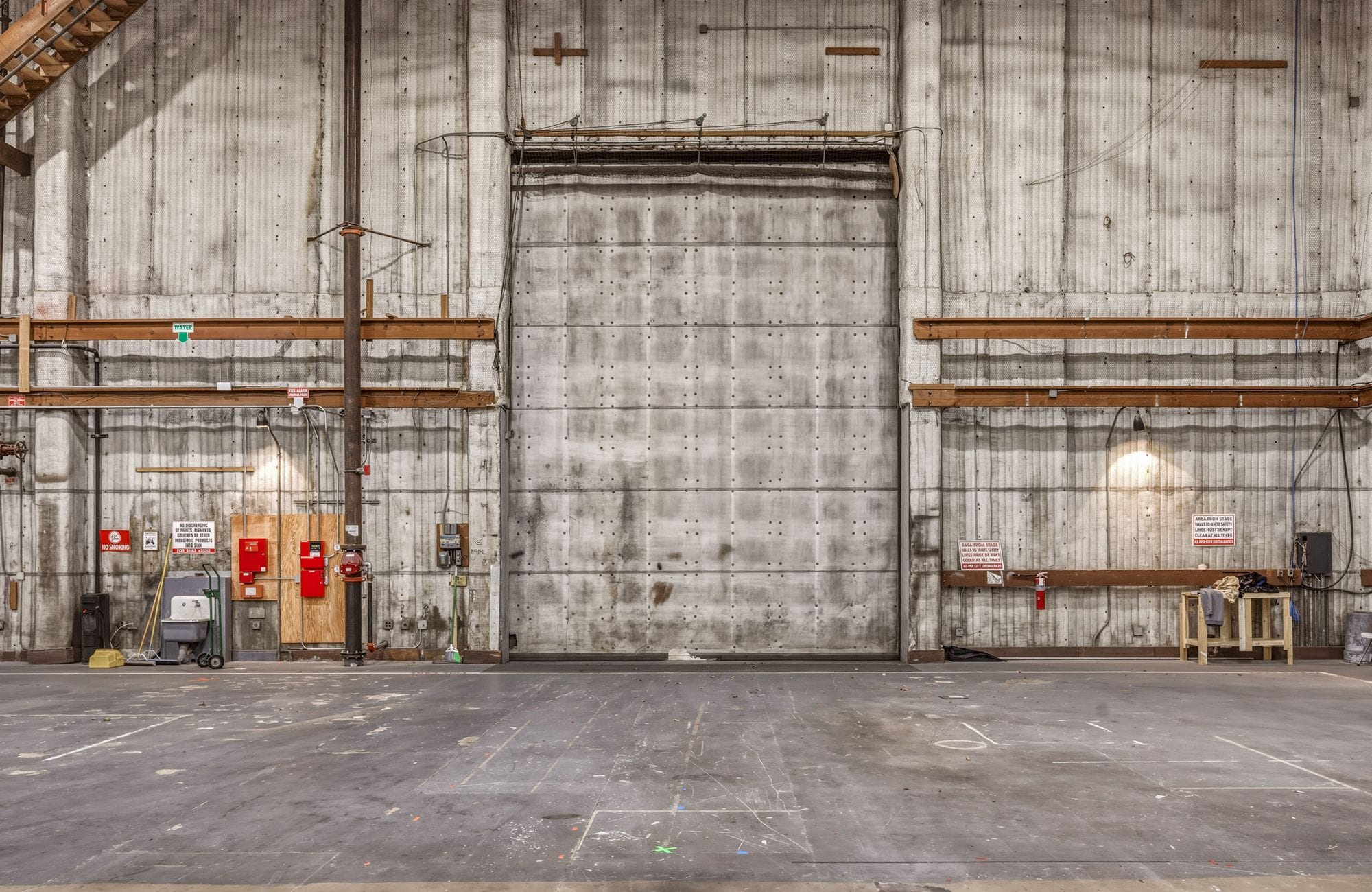 Interior of a large, weathered industrial building with a prominent gray roll-up door, concrete walls, and an empty floor.