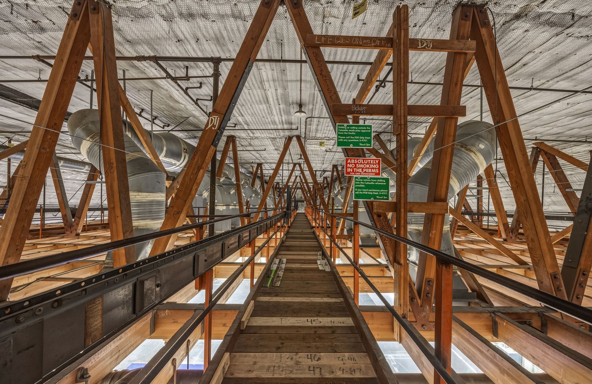 A long, elevated wooden and metal truss structure, like a catwalk, extending into an industrial space with large silver ventilation ducts overhead.