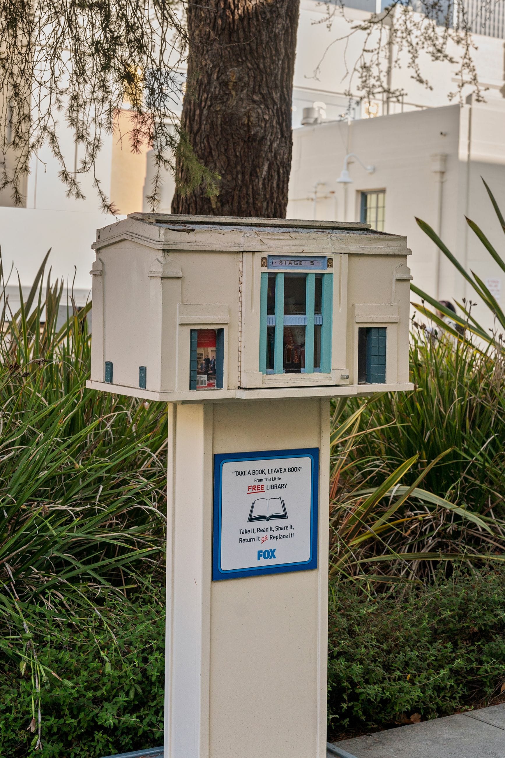 A miniature cream and teal building, labeled "STAGE 8," functions as a Little Free Library on a post. A sign below reads "Take a book, leave a book, FREE LIBRARY" and includes a Fox logo, with a tree and greenery.