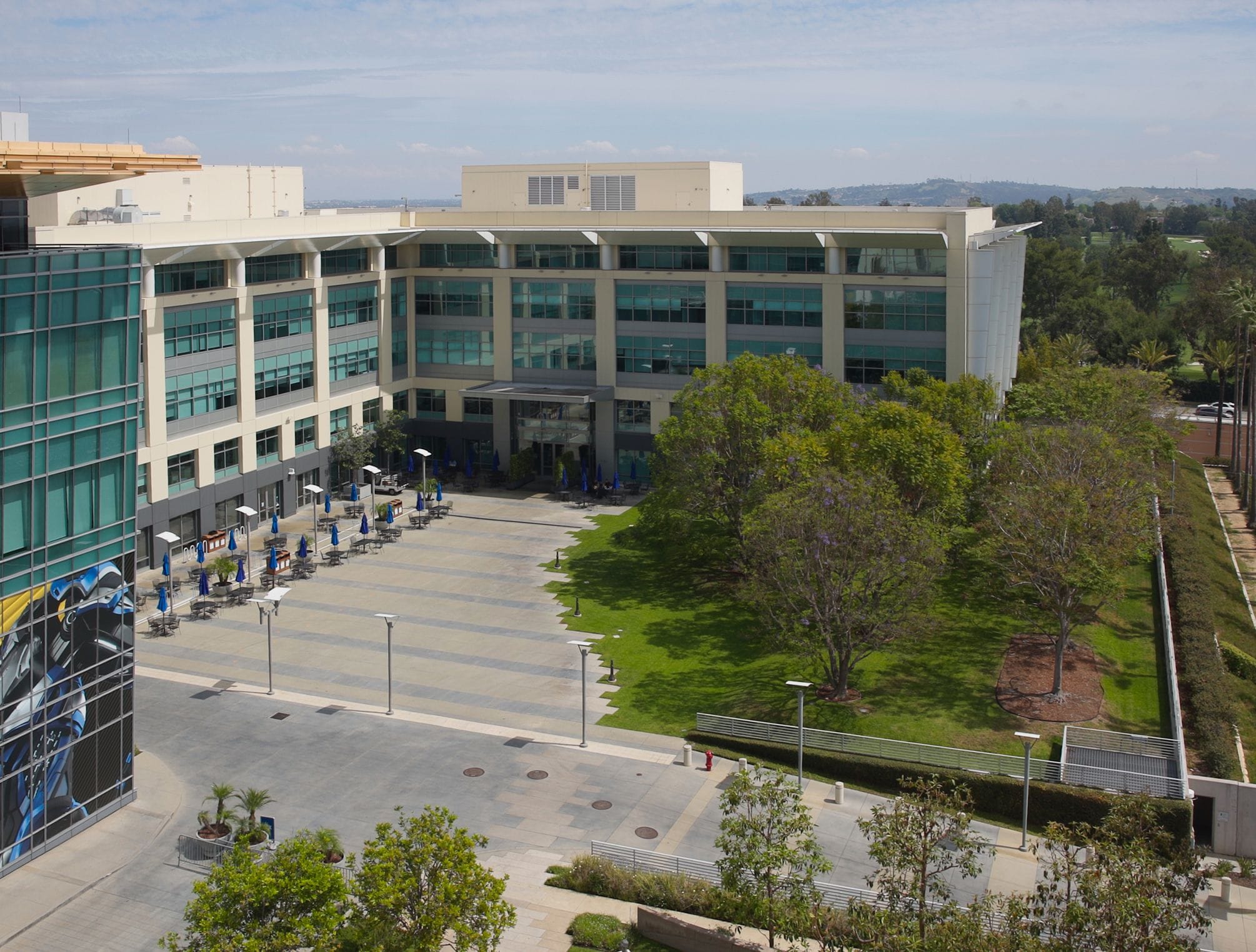 A modern office complex with an outdoor patio, green space, and a glass building displaying a large blue mural.