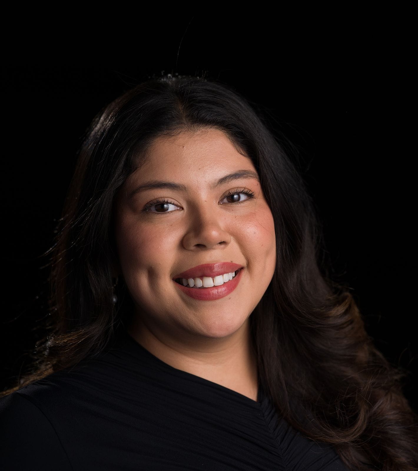 Headshot of a smiling woman with dark, wavy hair and dimples, wearing a black top against a black background.