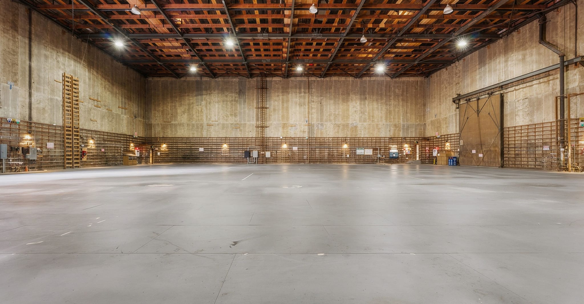 A large, empty concrete warehouse or sound stage with a high wood-beamed ceiling and overhead lights.