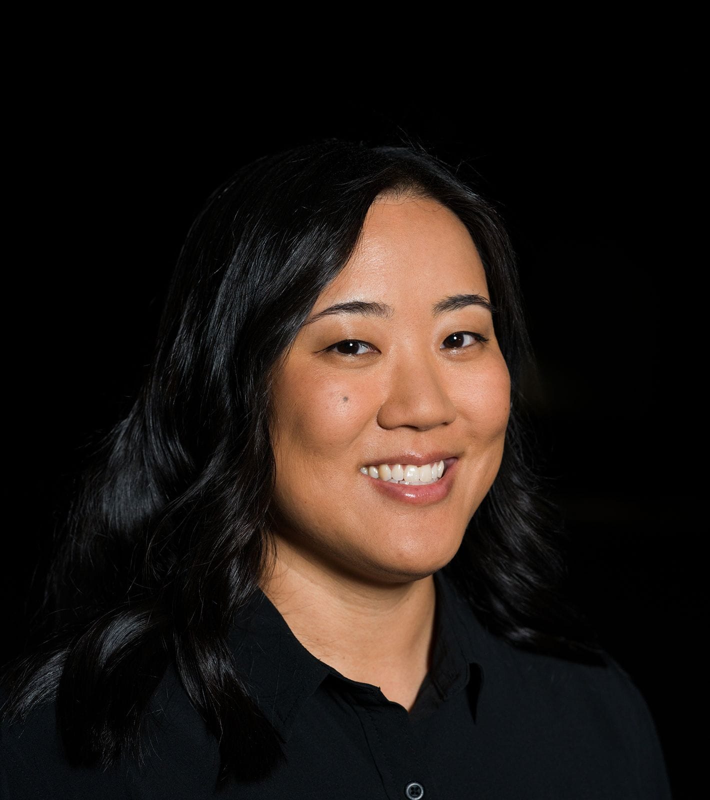 A smiling woman with dark hair wearing a black shirt against a black background.