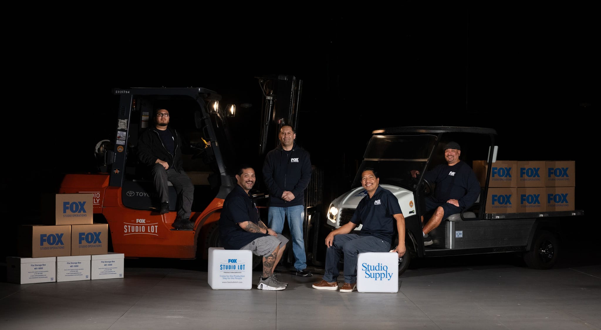 Five men pose with a forklift, a utility cart, and stacked boxes branded FOX, FOX Studio Lot, and Studio Supply.