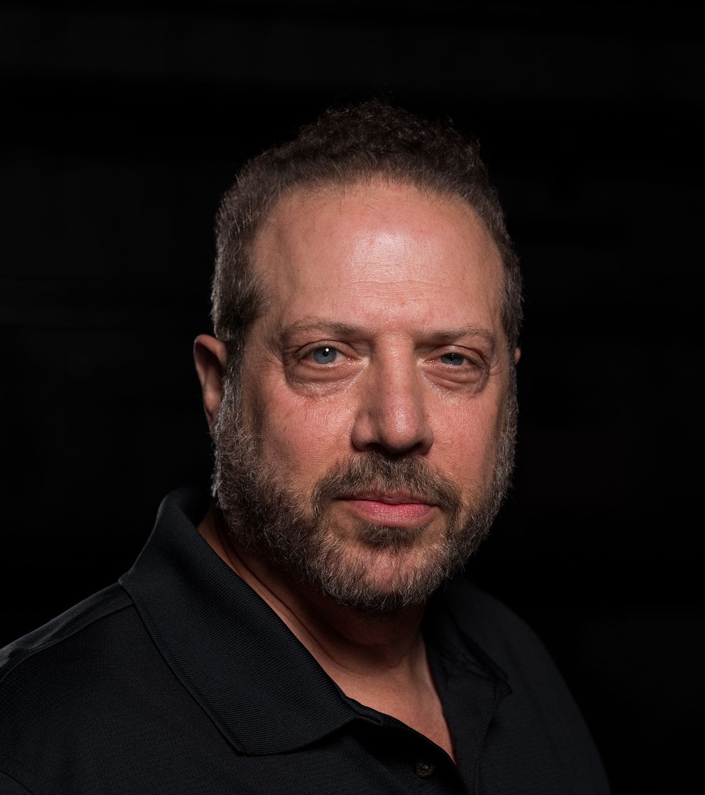 A middle-aged man with a beard and short dark hair wearing a black polo shirt looks directly forward against a dark background.