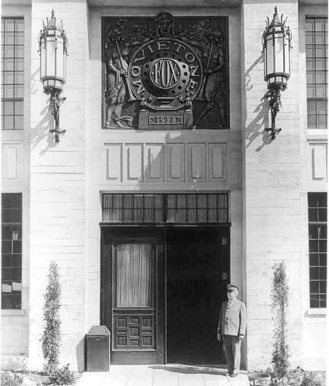 Black and white photo of the Fox Movietone building entrance, featuring a large 1928 plaque and a uniformed man.