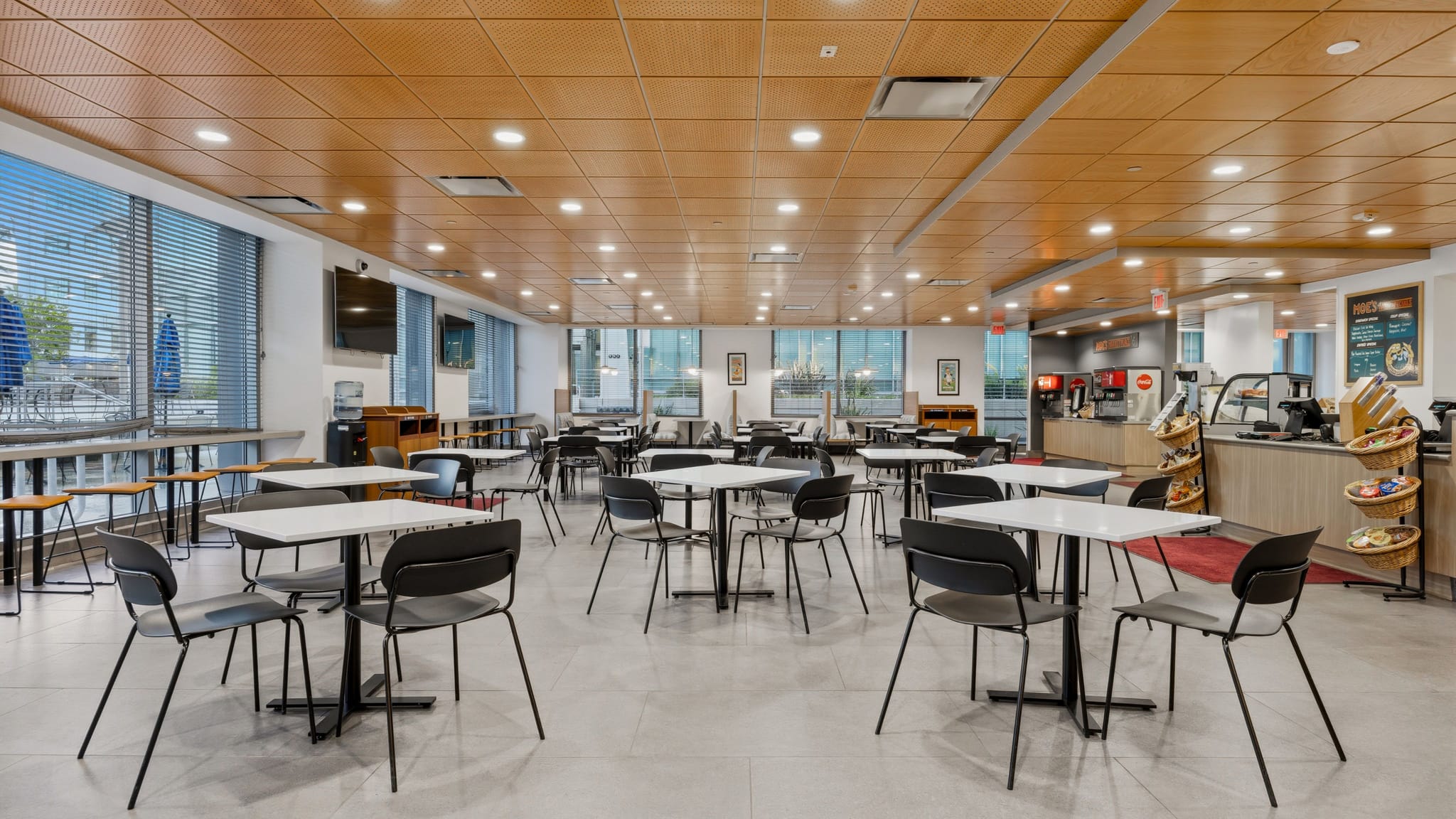 An empty, modern cafeteria with white square tables, black chairs, large windows, and a food service counter.