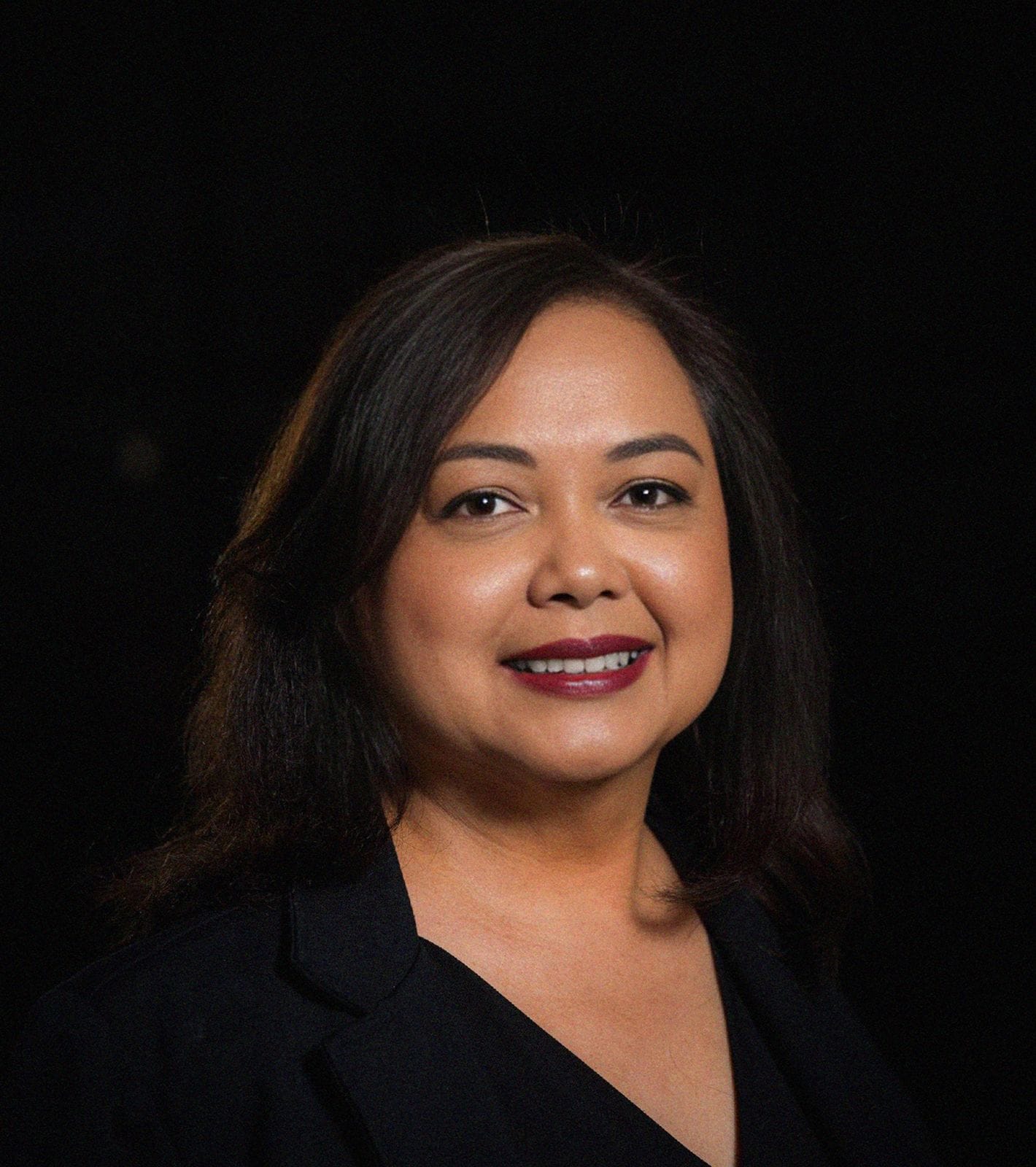 Smiling woman with dark hair, red lipstick, and a black blazer against a dark background.