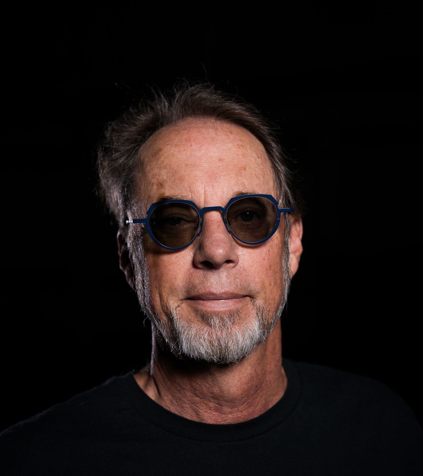 A man with a gray goatee and blue-framed sunglasses looks forward against a dark background.