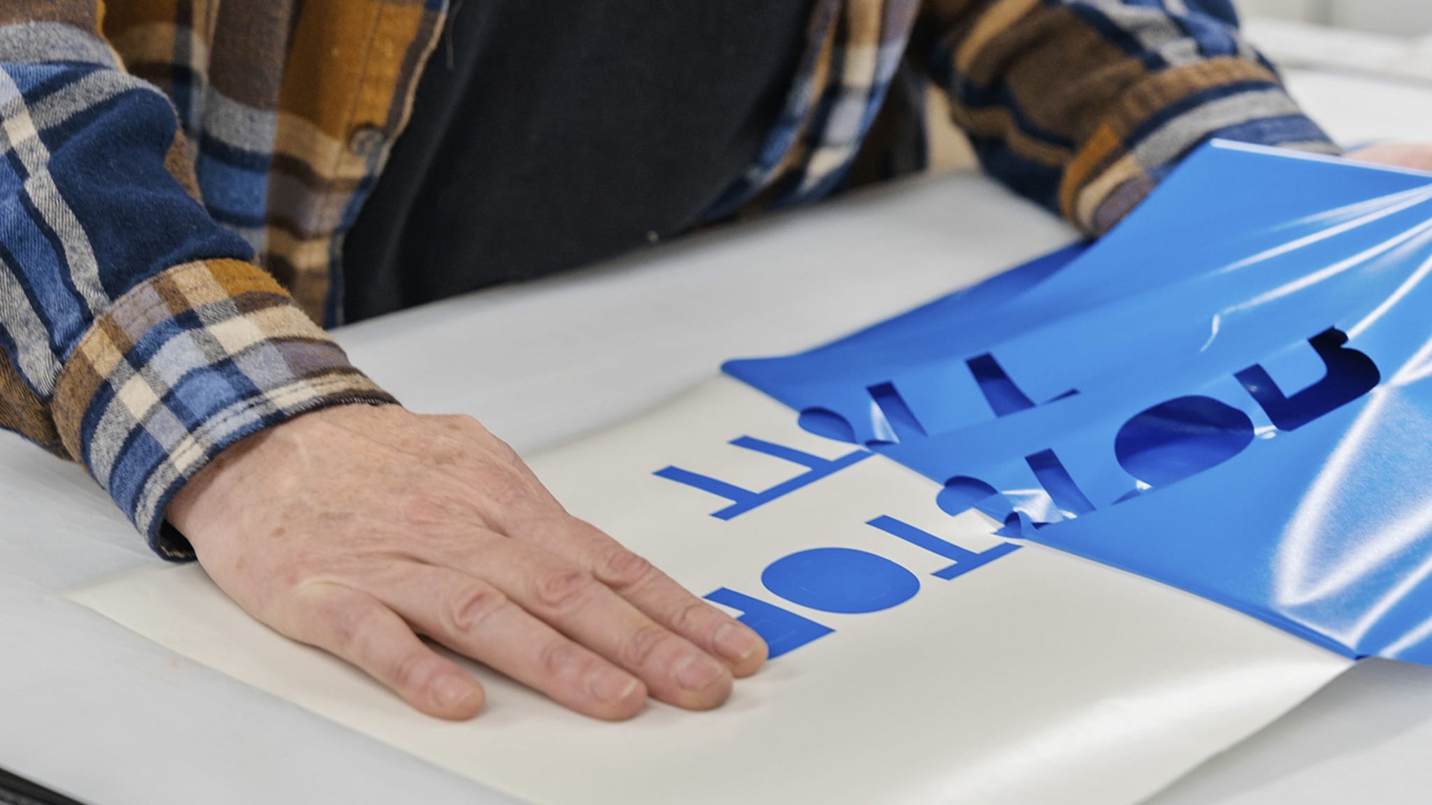 Hands peel blue vinyl revealing blue letters on a white surface.
