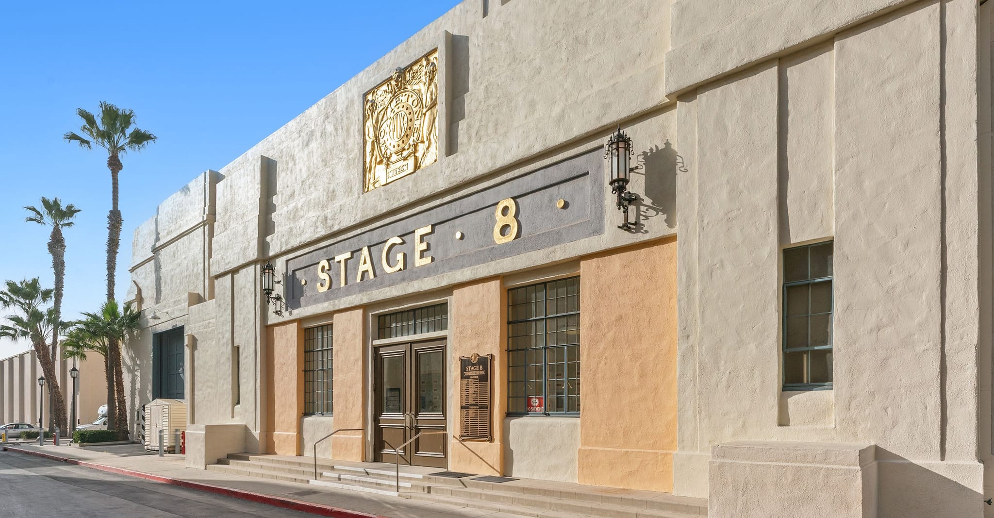 Art Deco building exterior with "STAGE 8" on its facade, a golden emblem, and palm trees.