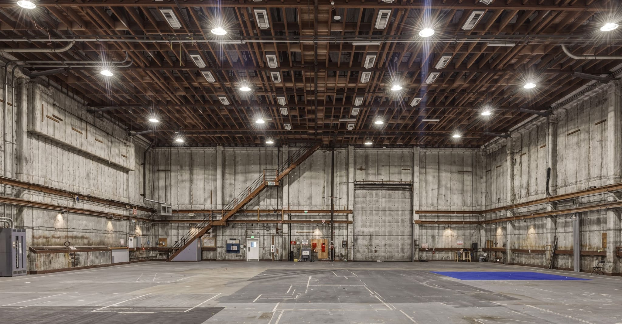 An empty, expansive industrial warehouse or sound stage with a high wooden ceiling, bright lights, and concrete walls.