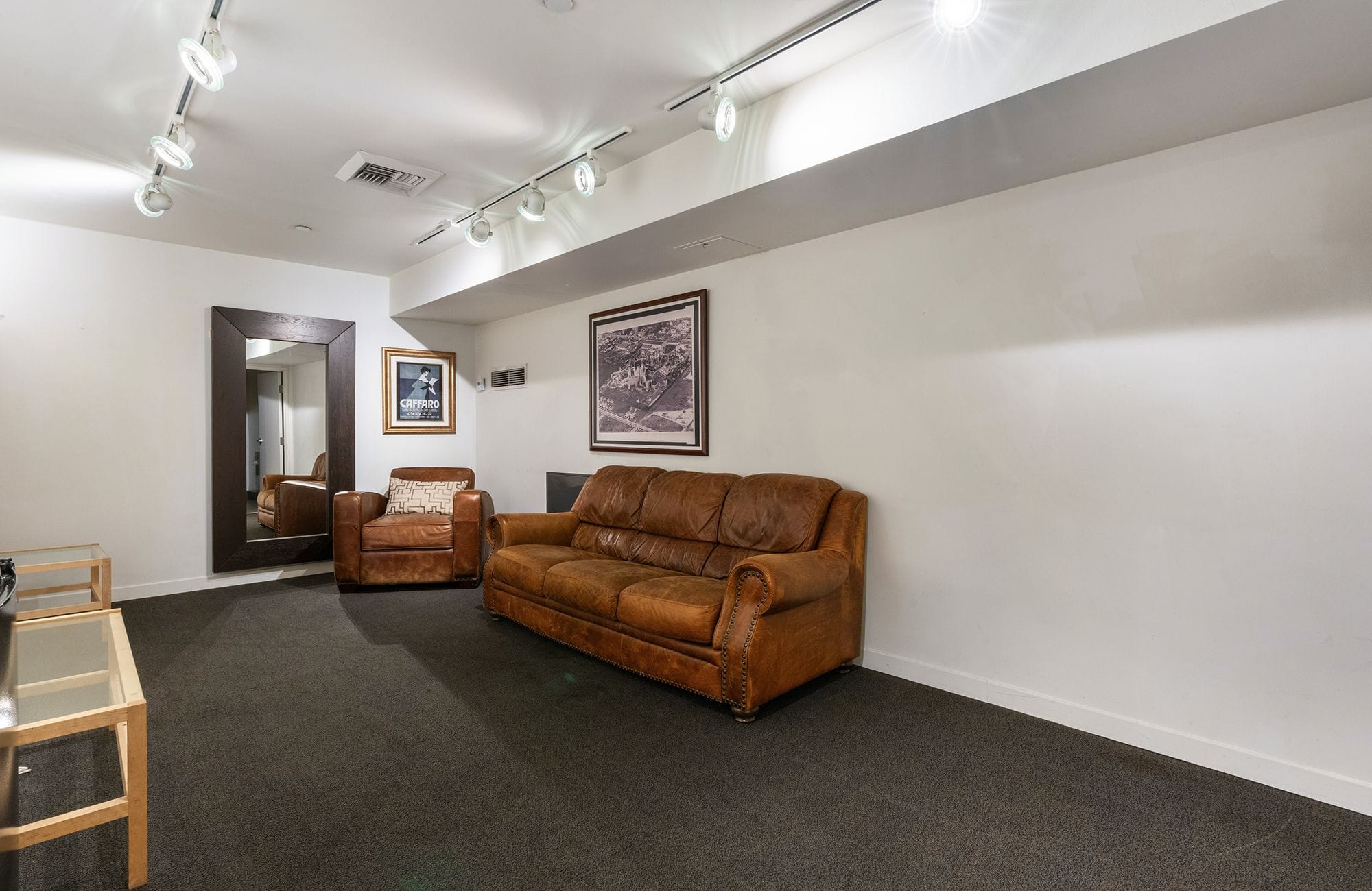 A room with white walls, dark carpet, brown leather furniture, a large mirror, and track lighting.